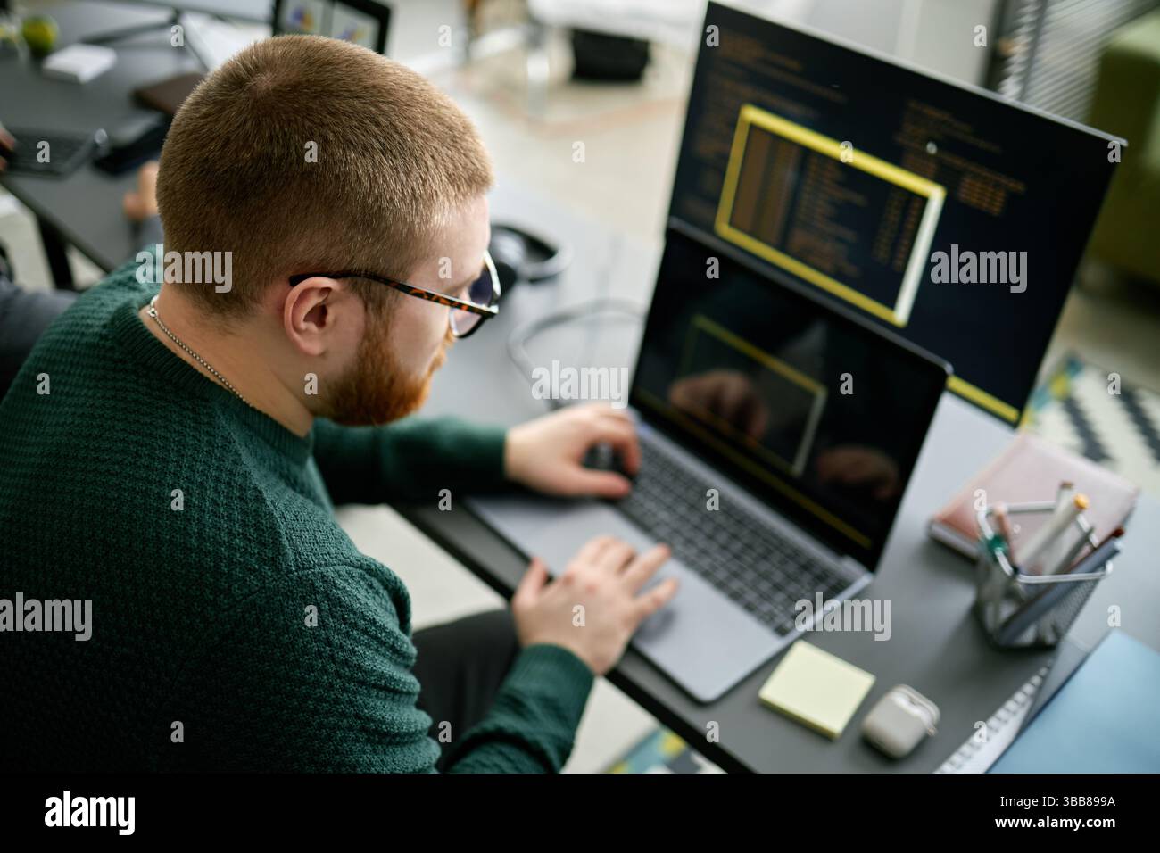 Coder working on project with two monitors and a laptop, immersed in code. Office environment showing programmer focused on software development Stock Photo