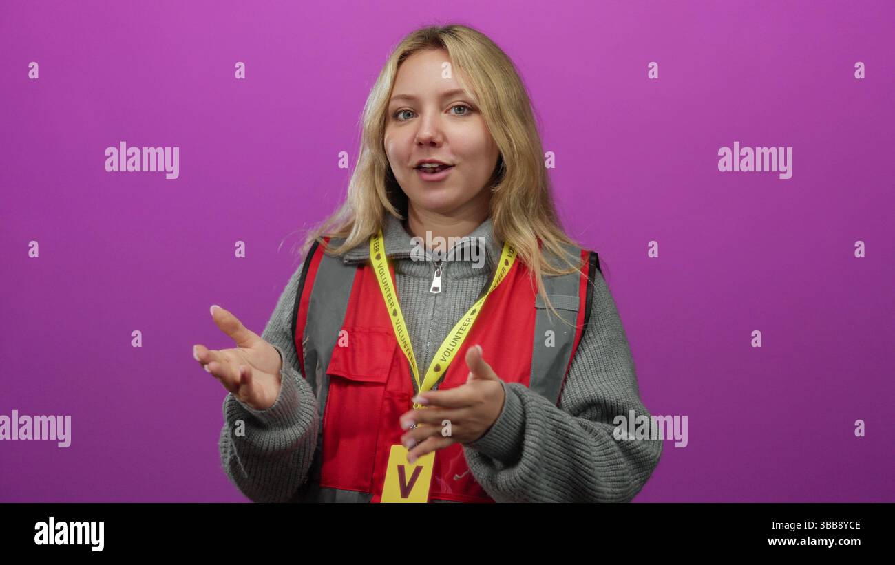 Woman volunteer with blonde hair expressing emotions against a vibrant pink background wearing a red vest and yellow lanyard. Stock Photo