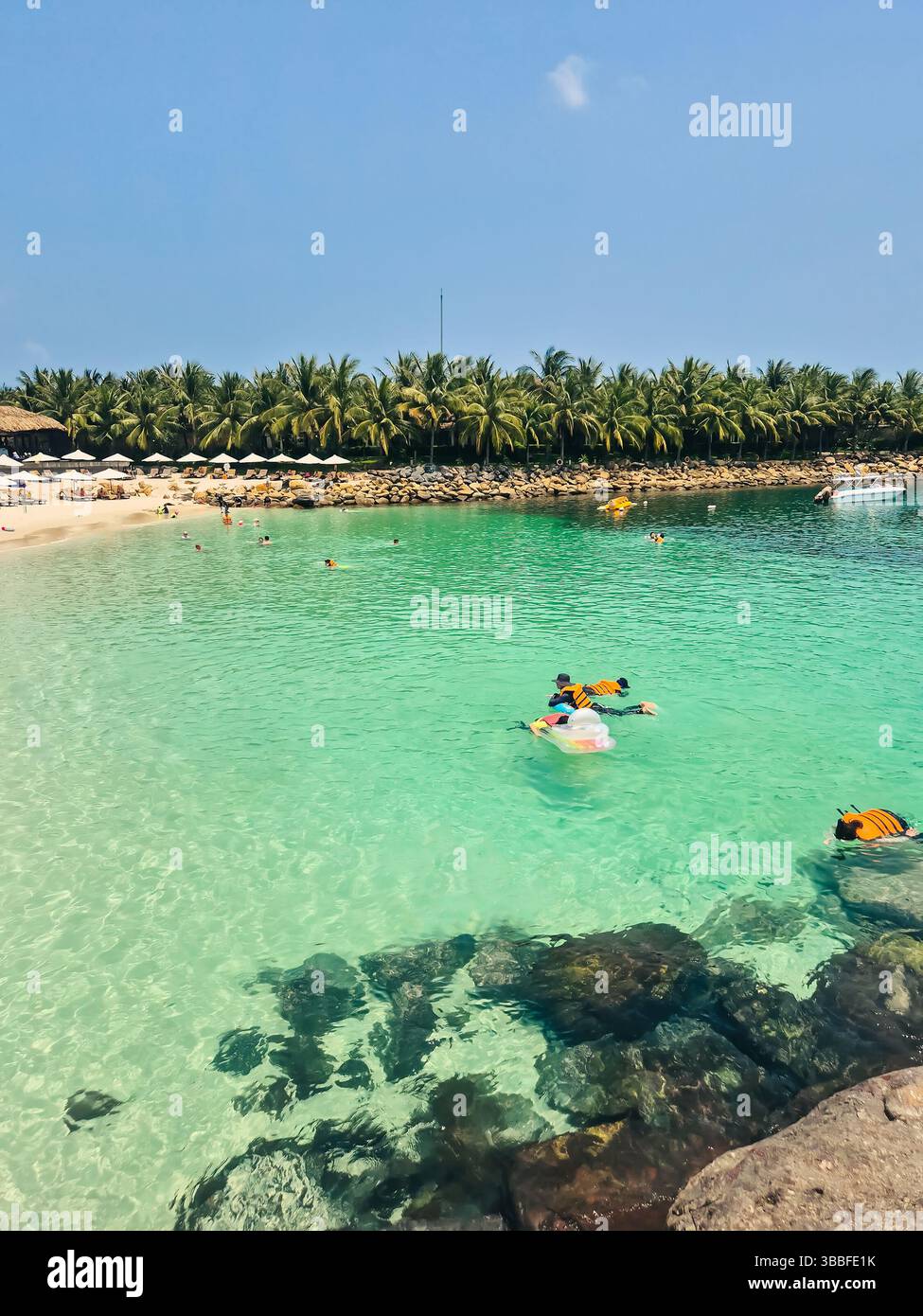 Tropical beach scene in Vietnam with swimmers in life jackets, crystal clear water, horizon view, white sand, rocks and palm trees. Nha Trang coastlin Stock Photo