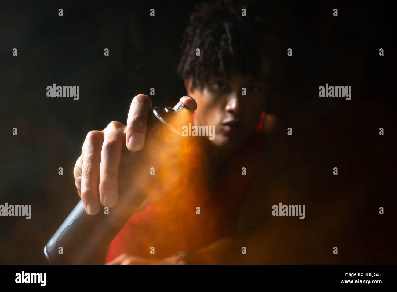Holding black spray paint can, African American male releasing bright orange mist in dark studio Stock Photo