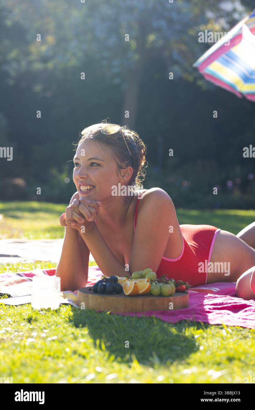 Lying woman wearing red swimsuit on towel in garden, with fruit board and drink under umbrella Stock Photo
