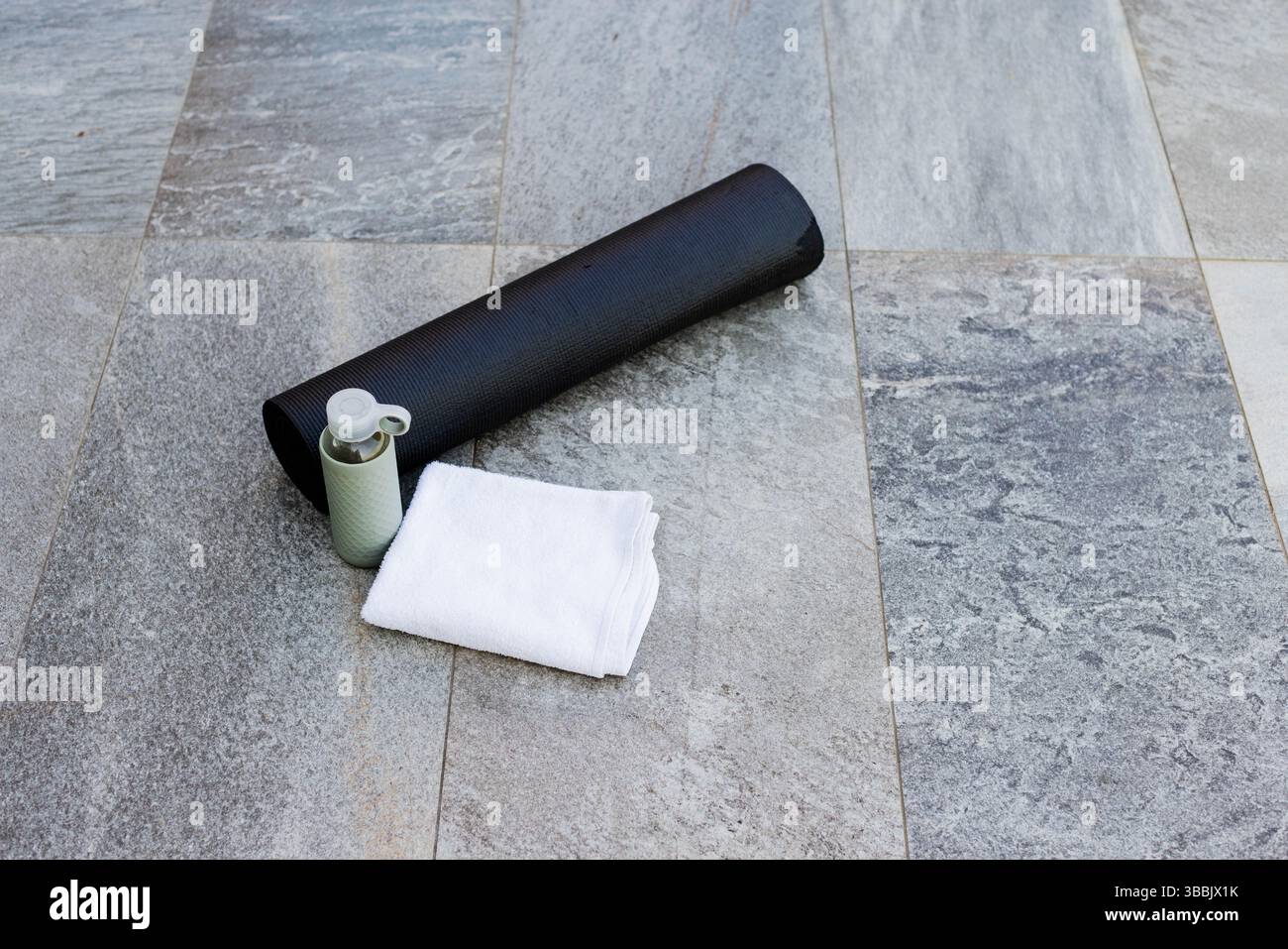 Rolled black exercise mat lying diagonally on grey patio with white towel and green water bottle Stock Photo