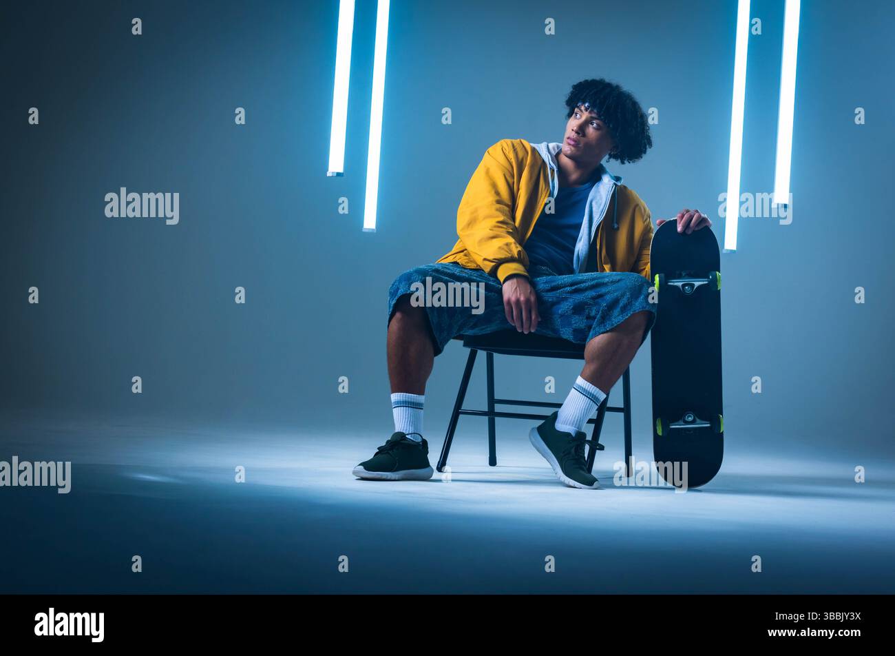 Sitting African American man wearing jacket and shorts on chair in studio, holding skateboard Stock Photo