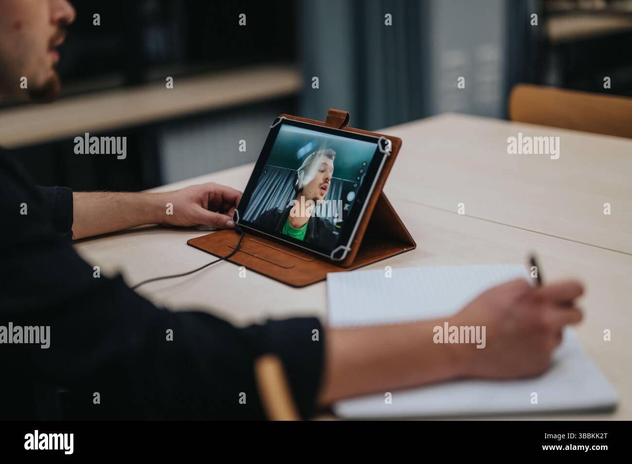 Young man participating in an online video call while taking notes on a notepad at a table Stock Photo