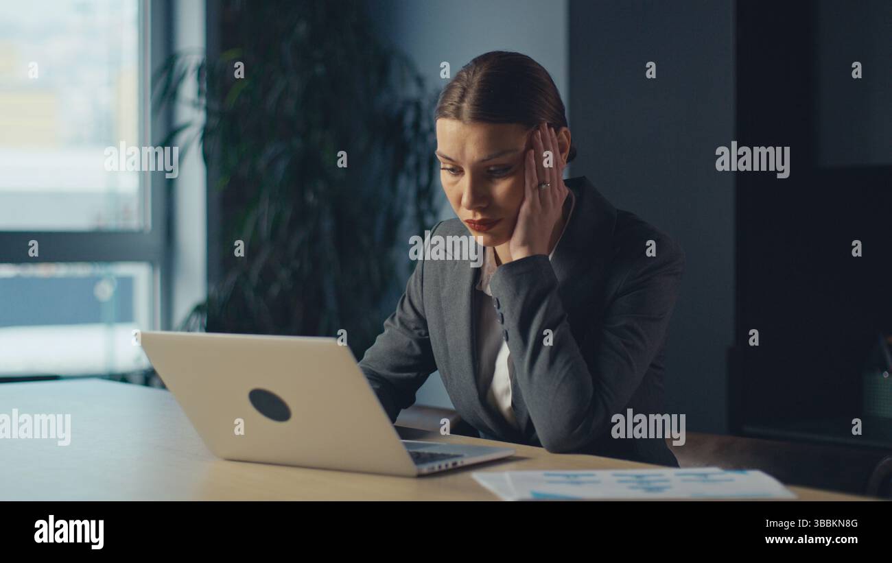 Stressed businesswoman in a formal suit sitting at desk and looking at laptop screen in low light office. Concept of workplace anxiety, corporate Stock Photo