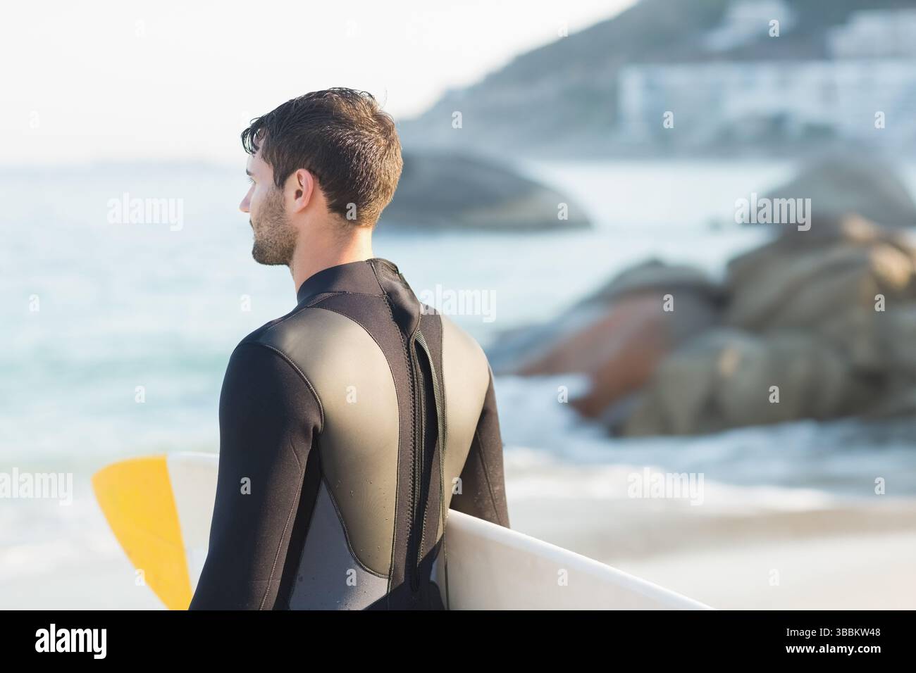 White surfboard resting on sandy shoreline facing calm ocean with rocky boulders under clear sky Stock Photo