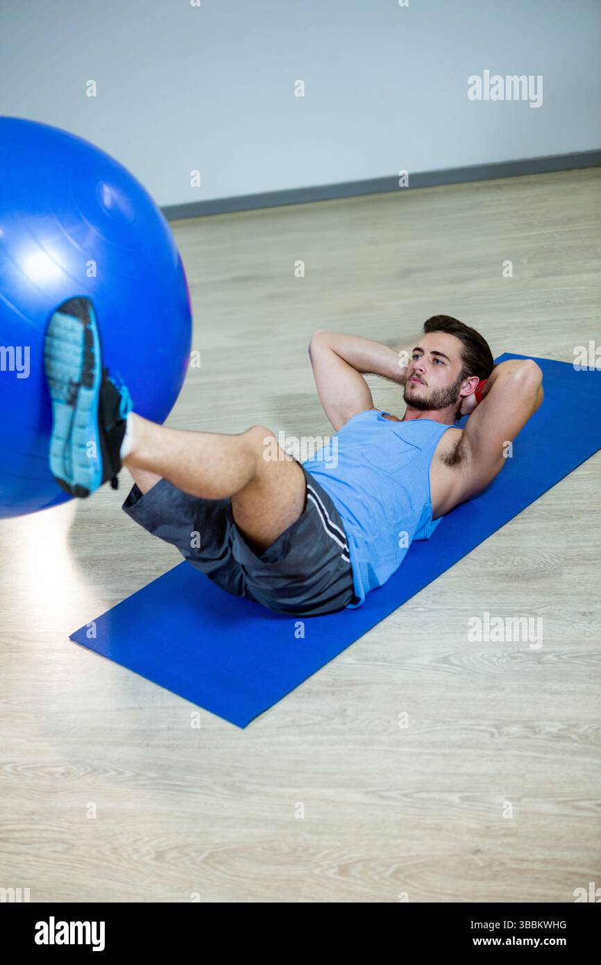 Young man lying on blue yoga mat at gym, doing core workout with ball, copy space Stock Photo