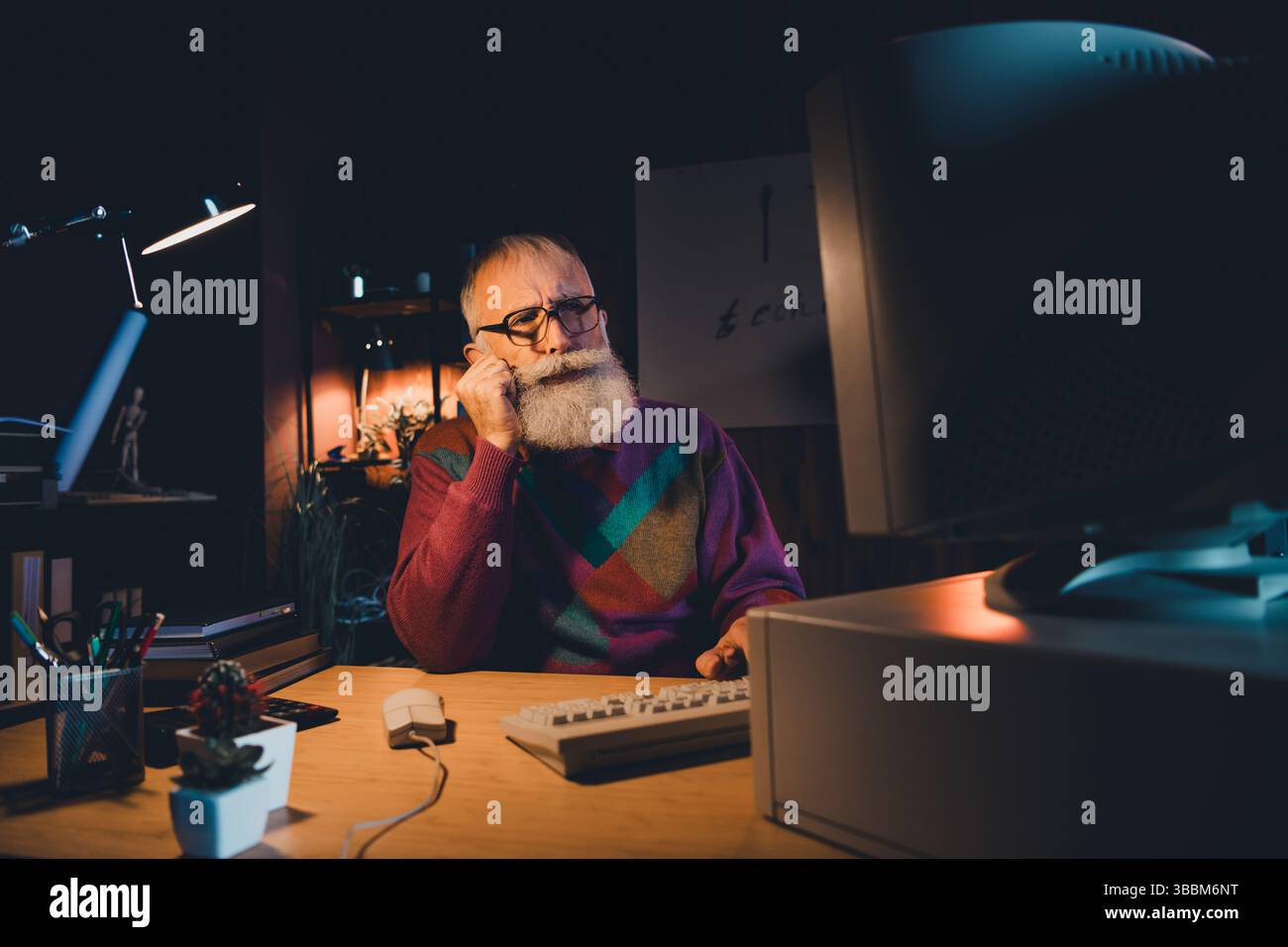 Elderly programmer working late at home office desk, wearing colorful sweater, focused on computer coding development Stock Photo