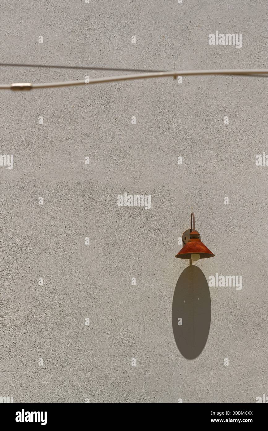 A white stucco or rendered wall with a single lamp with a rusty shade. A strong shadow cast below. Stock Photo