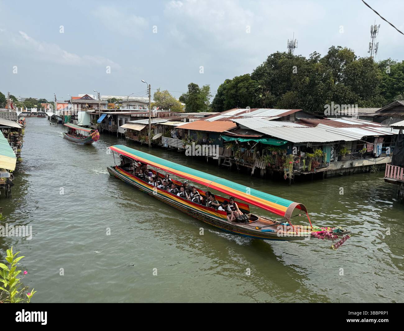 A colorful longtail boat cruises past riverside homes in a Thai canal, capturing daily life along Bangkok’s historic waterways - Smartphone Captured Stock Image