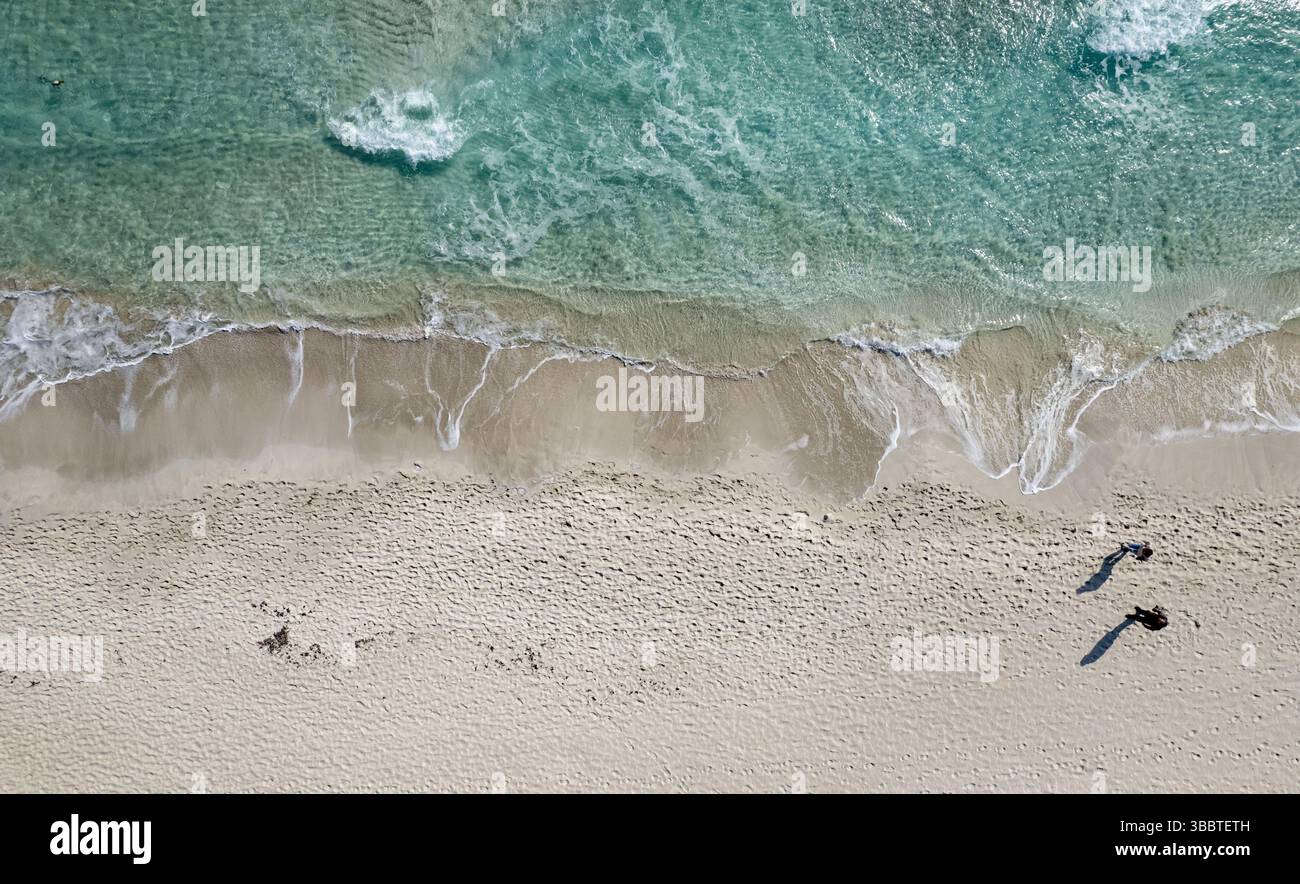 Drone Aerial view of waves washing ashore on a white sand beach with two tourists walking, enjoying their vacation. Ayia Napa Cyprus Stock Photo