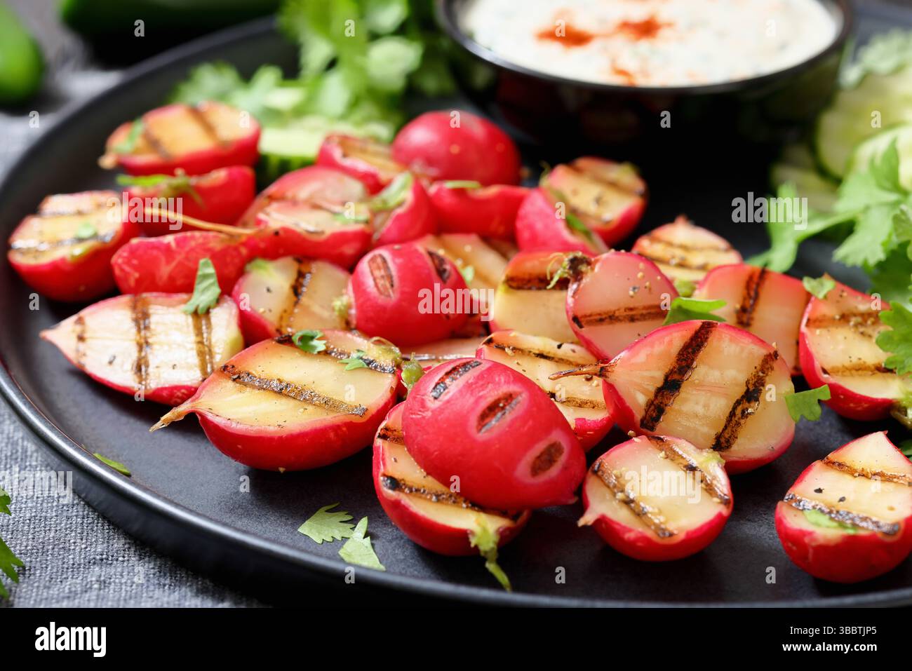 Grilled radishes with cucumber slices and sour cream sauce served on a dark grey plate, close-up, real photo, not AI generated Stock Photo