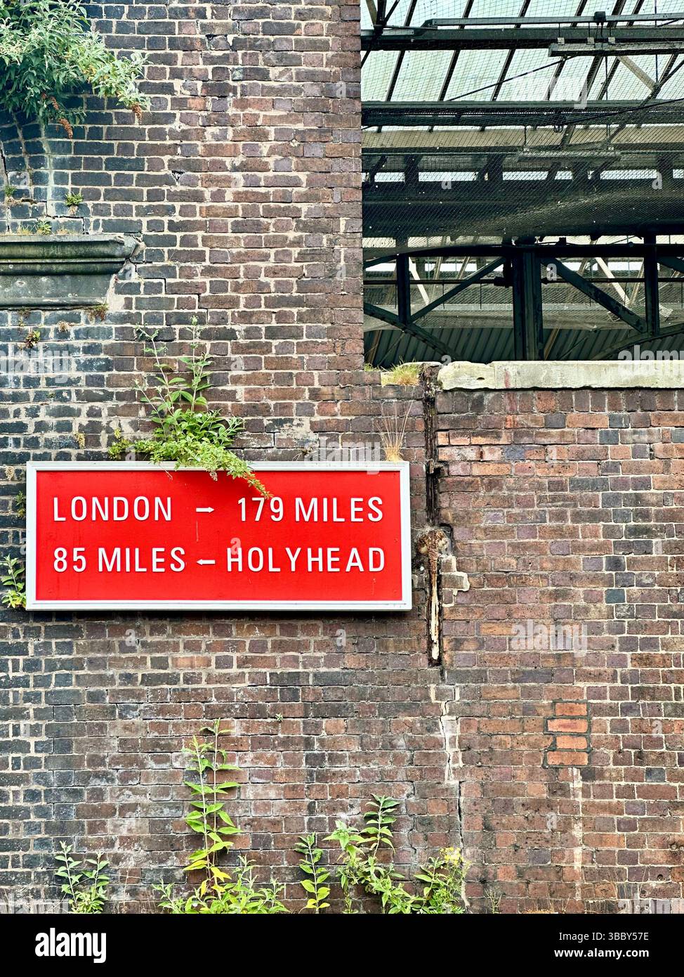 Vintage sign at Chester train station showing distances to London and Holyhead. Worn brick wall with small plants. 17 May 2025. - Smartphone Captured Stock Image