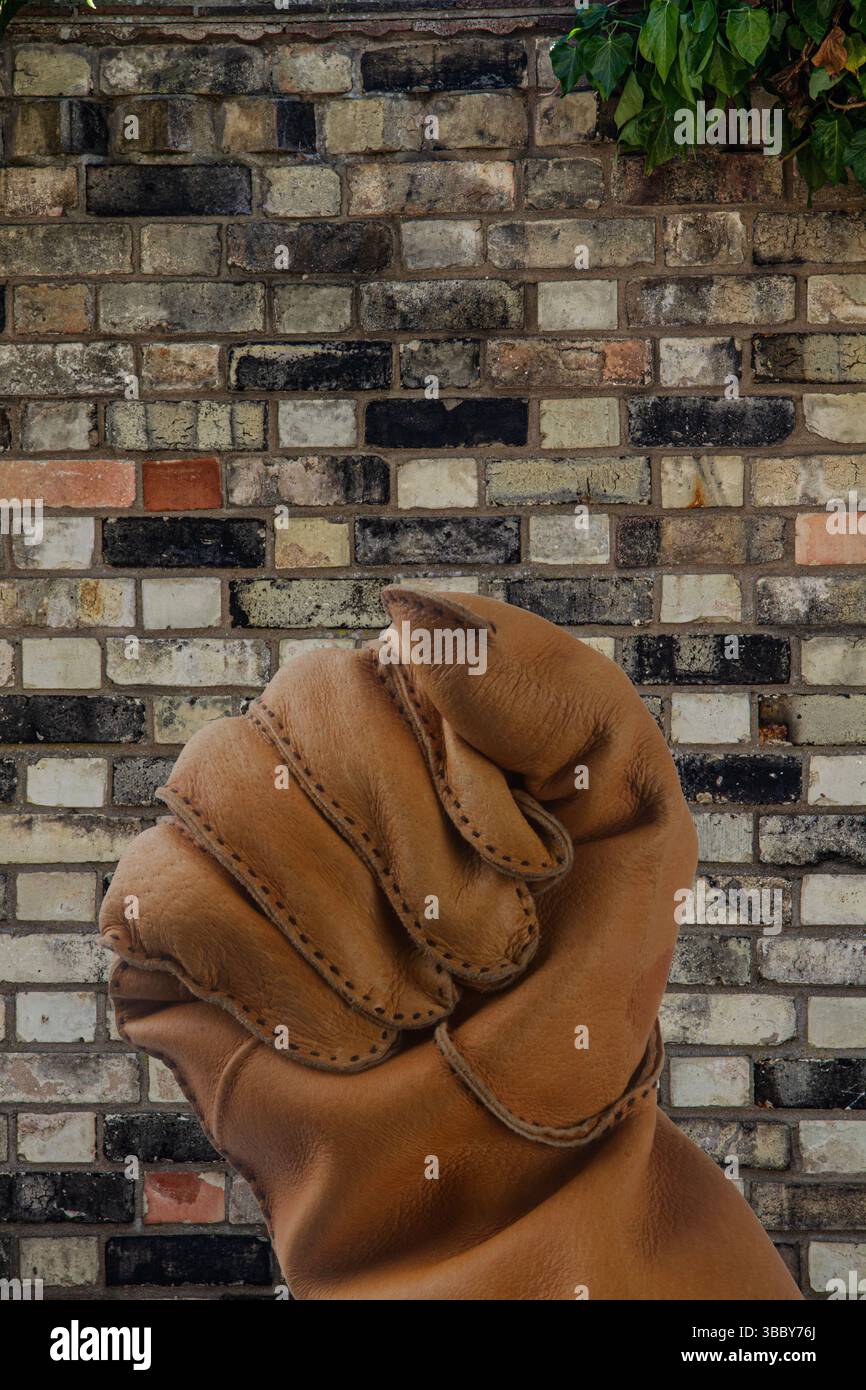 A held up gesture of a clenched fist in a light brown heavy leather glove against a background of a multicoloured Victorian ancient brick wall Stock Photo