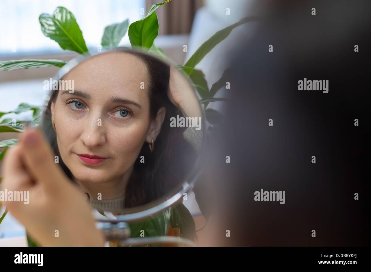 Woman looking at herself through a small round mirror, A woman looks thoughtfully at her reflection in a handheld round mirror, surrounded by plants. Stock Photo