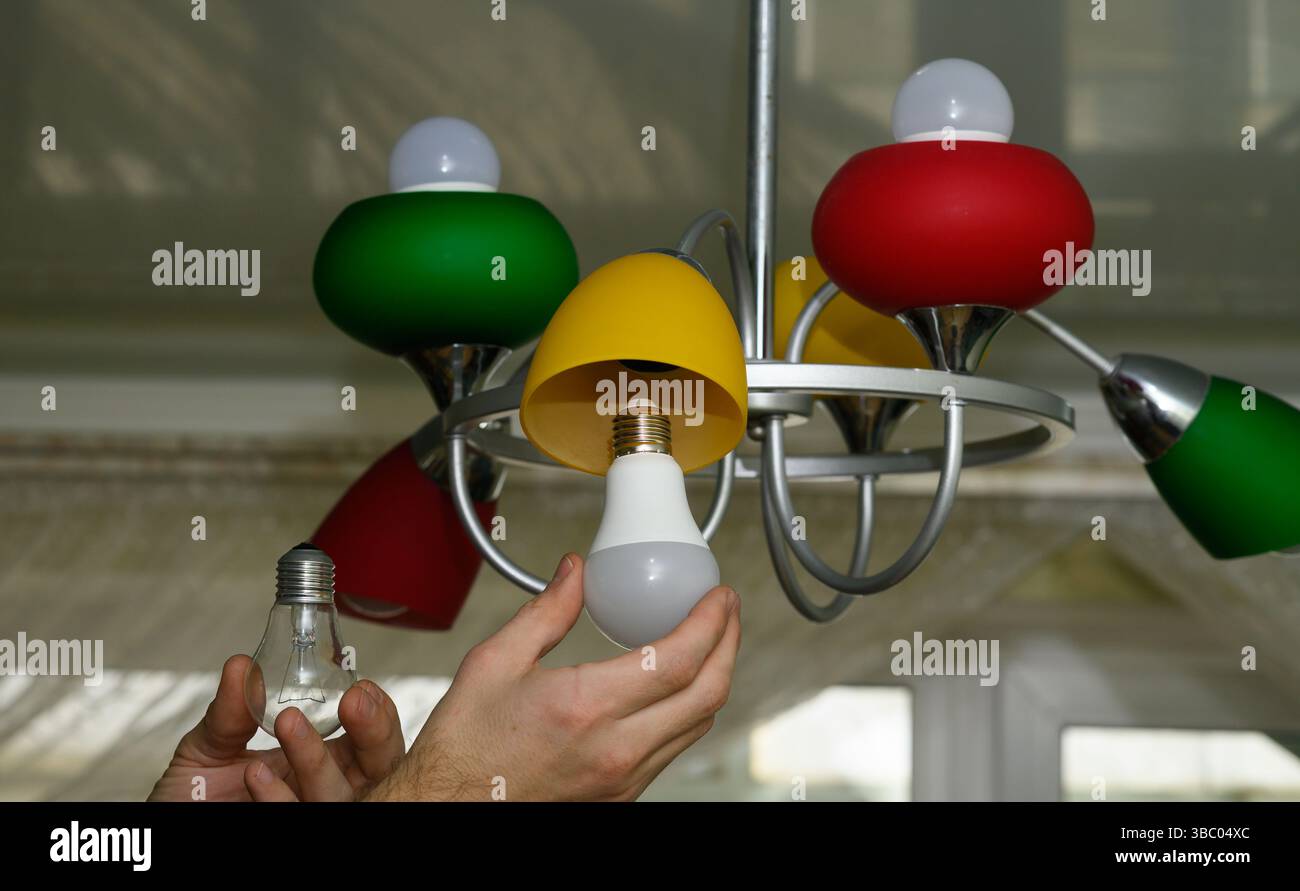 Male hands compare classic bulb with LED alternative while replacing lamp. Lamp highlights contrast between outdated energy use and efficient green so Stock Photo