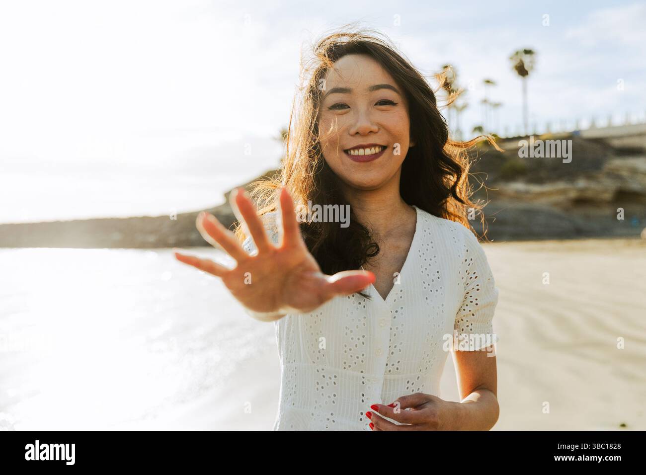 A woman is smiling and pointing her index finger at the camera. She is wearing a white shirt and is standing on a beach Stock Photo