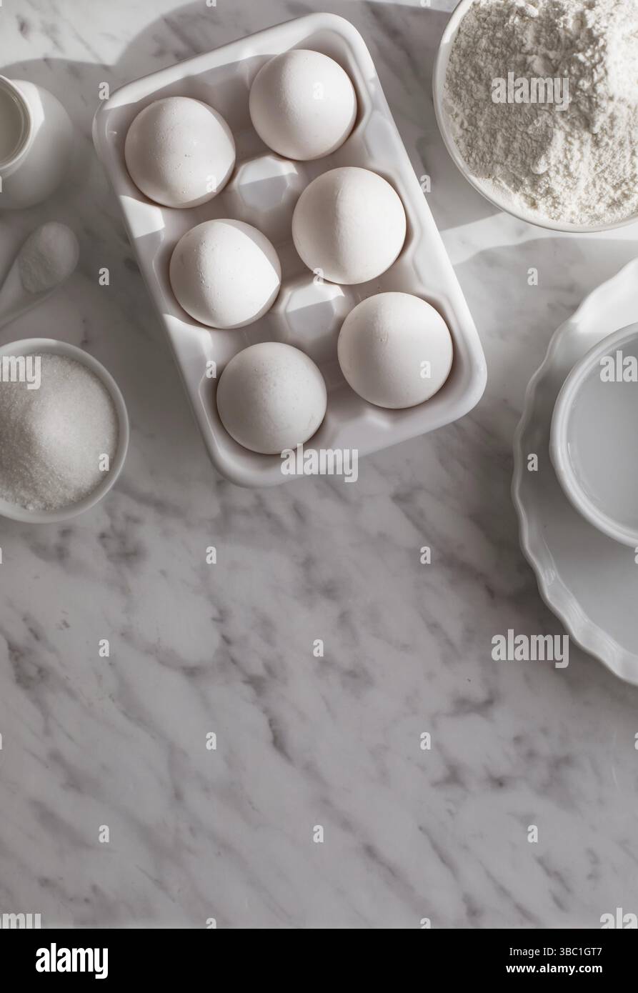Top view ceramic tray with eggs placed near bowl of flour and sugar. White utensils with white ingredients on white surface. Copy space Stock Photo