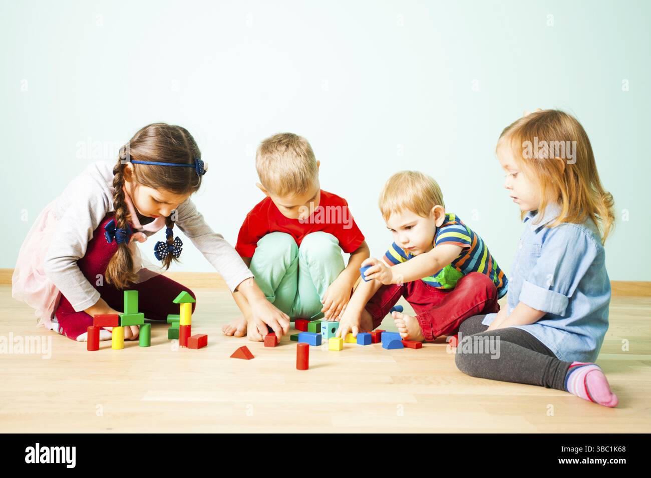 Kids of various age play with colorful blocks sitting on a wooden floor ...