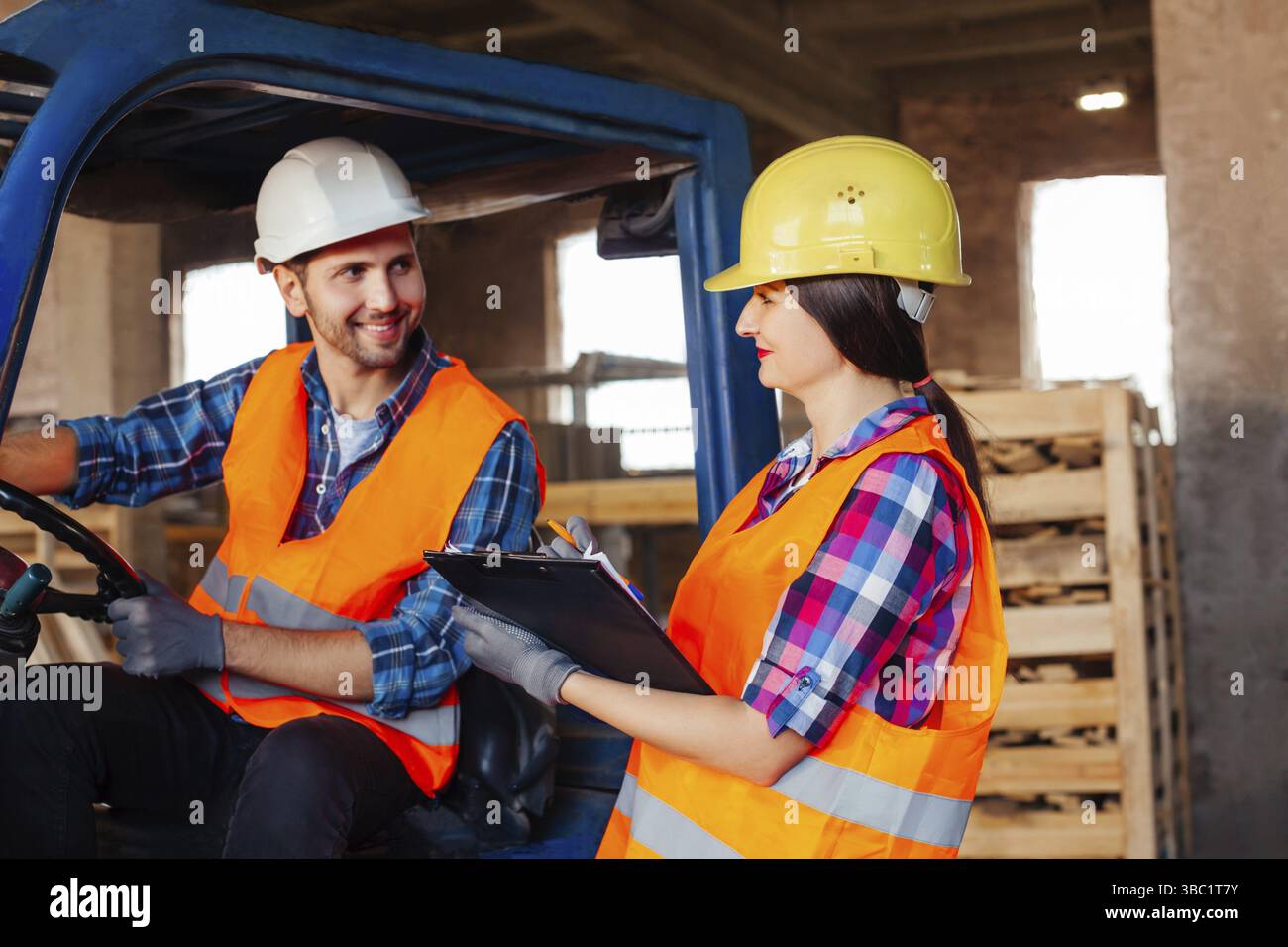 Male forklift driver sitting in a truck, female factory worker standing near, both discussing working issues. Warehouse workers in hardhats and orange Stock Photo