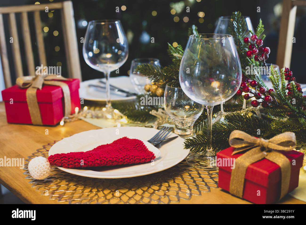 Christmas table setting with red crocheted Santa hat for cutlery on plates, festive decorations with fir tree branches. Christmas and New Year holiday Stock Photo