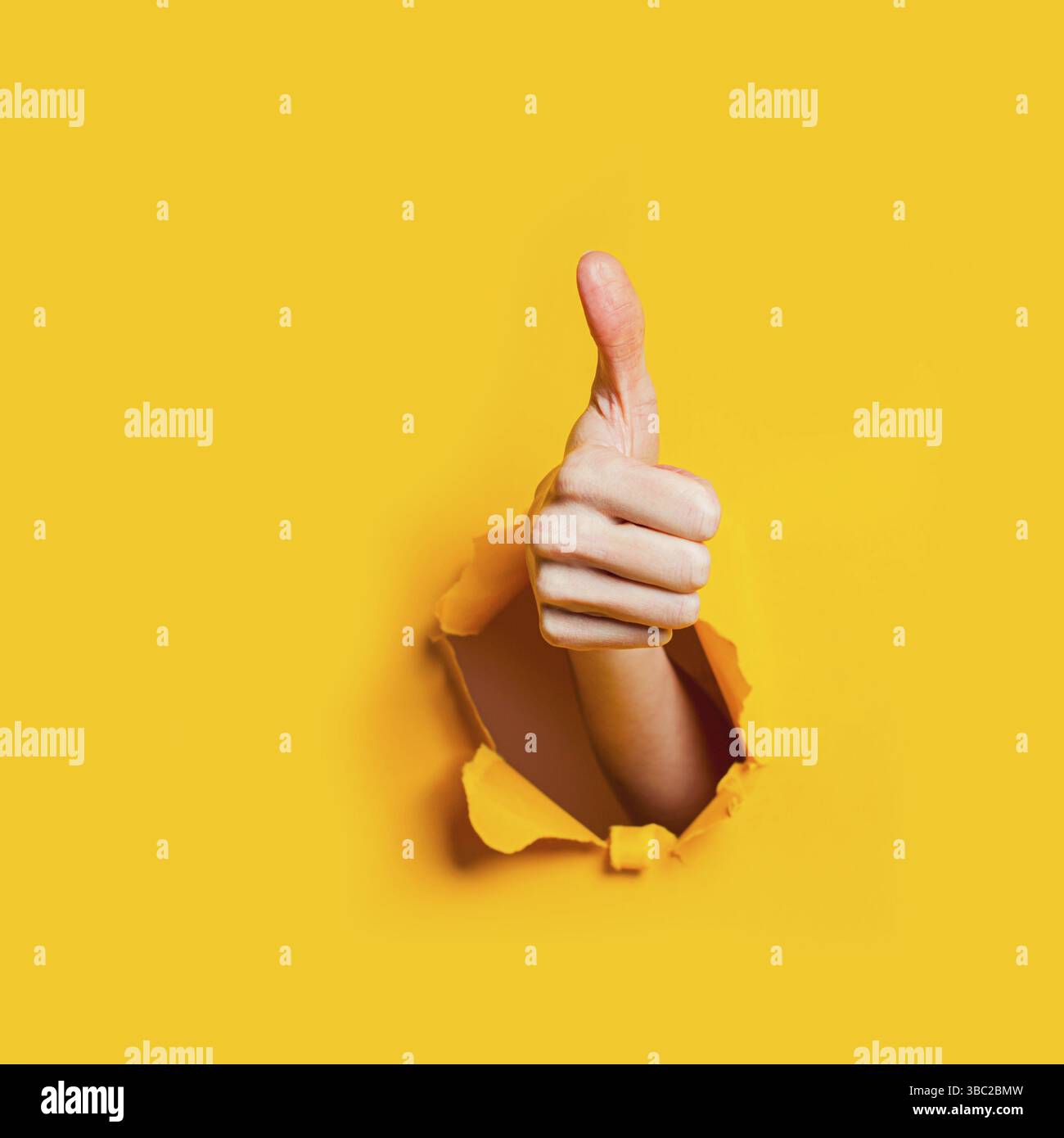 Hand showing a thumb up sign through a ripped hole in yellow paper ...