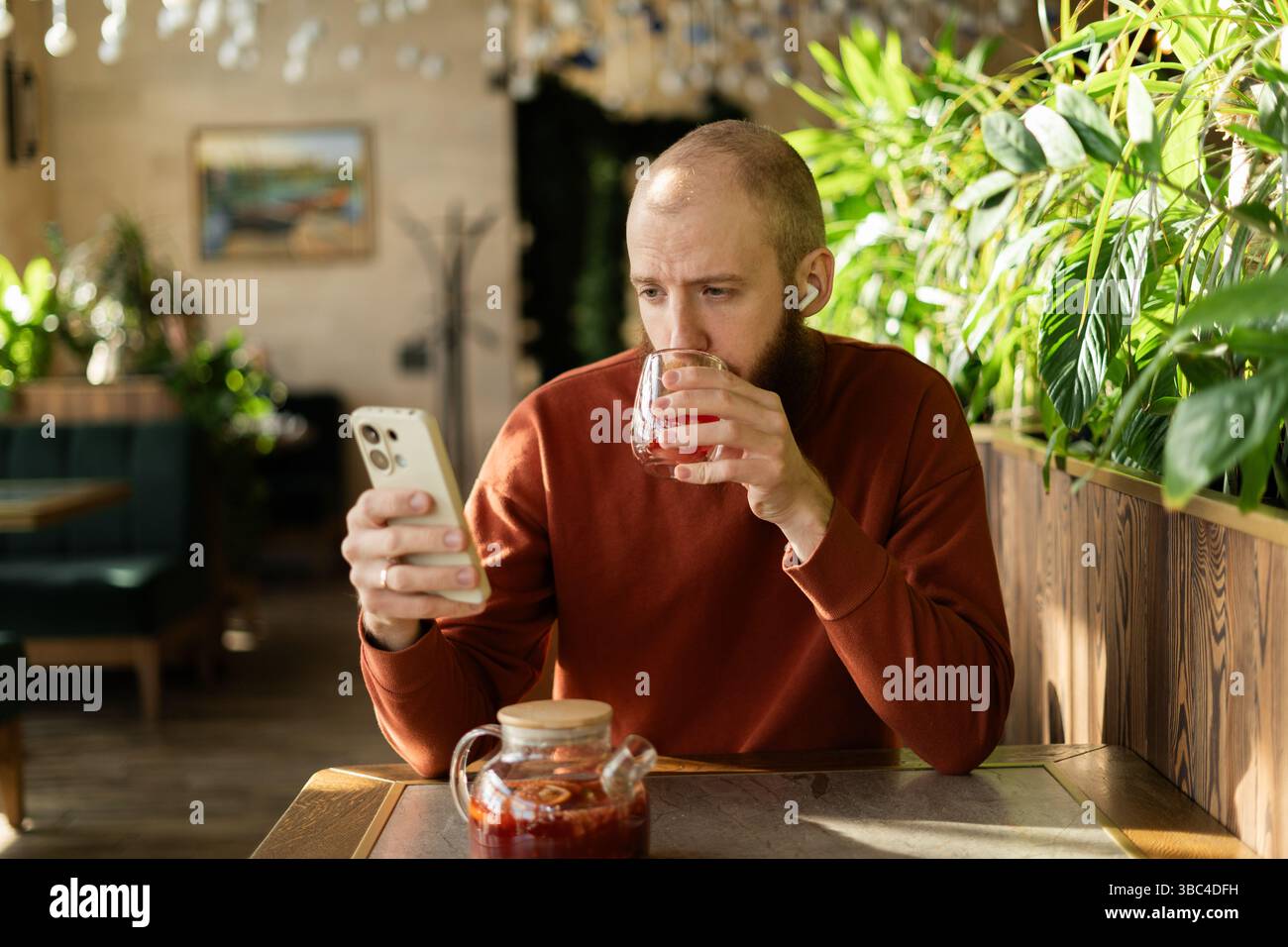Bearded handsome man drinking tea, reading news and social media using mobile phone at green cafe Stock Photo