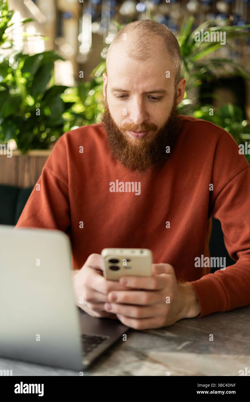 Millennial man using cellphone for chatting while sitting near laptop computer in office Stock Photo