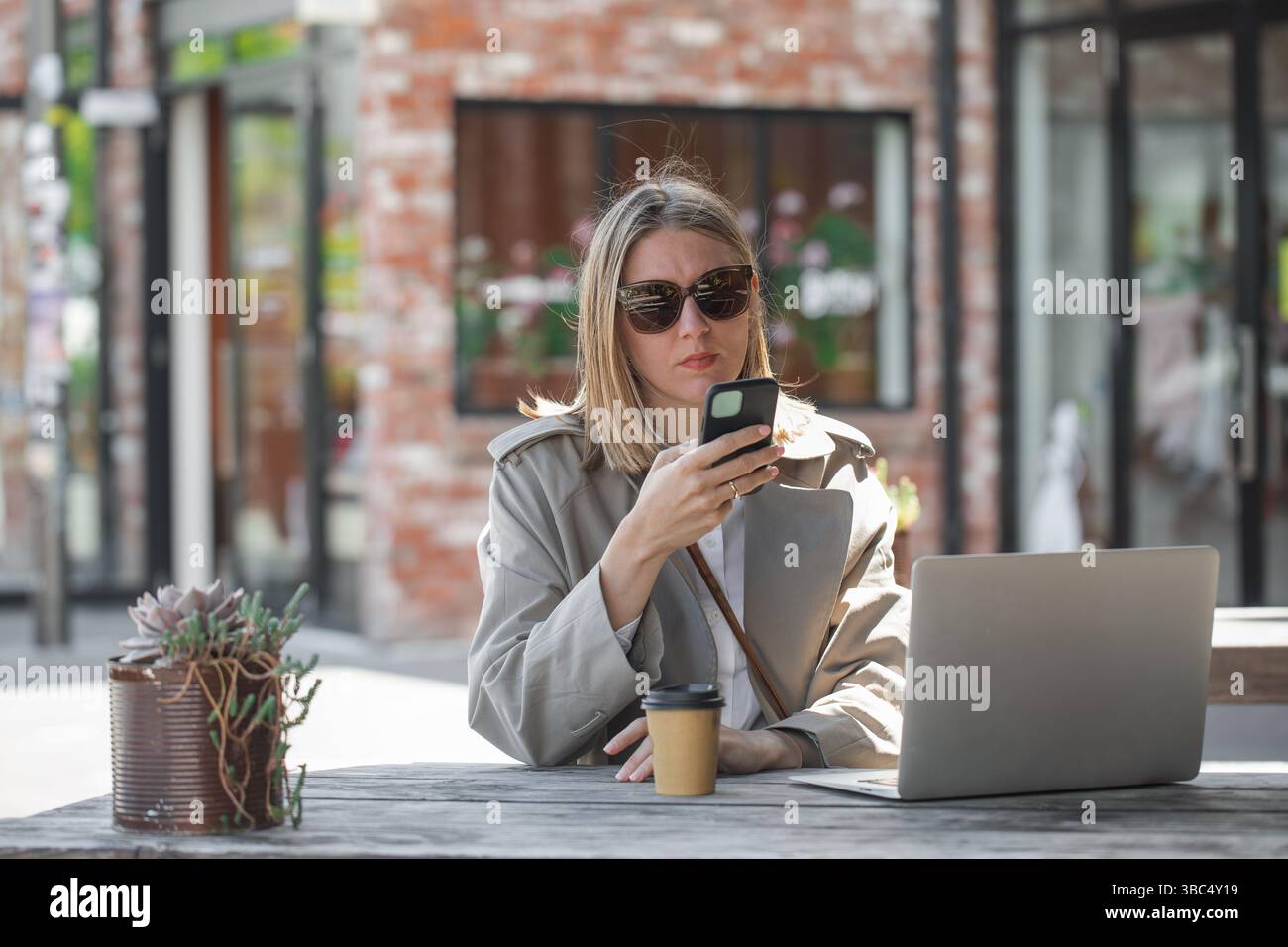 Young woman working using phone and laptop sitting on cafe veranda. Remote work, businesswoman Stock Photo