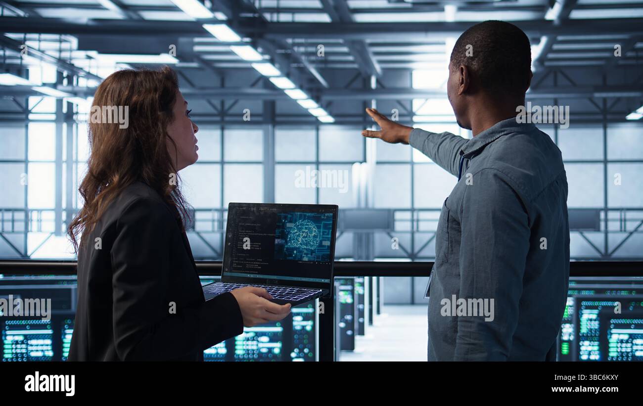 AI technology used by data center software developers reviewing equipment diagnostics. Programmers coding on laptop using artificial intelligence, adjusting server farm configurations, camera B Stock Photo