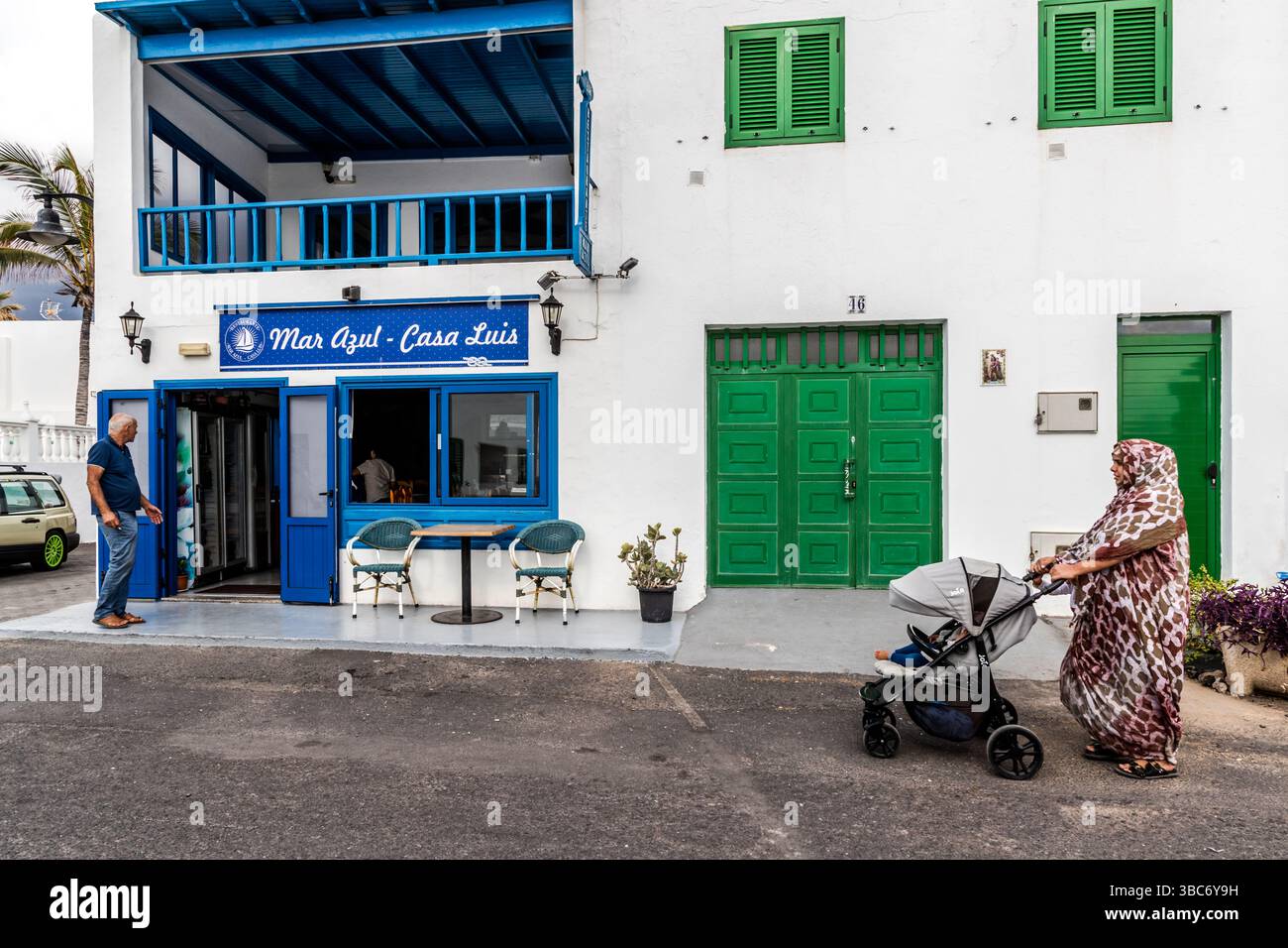 El Golfo on Lanzarote: A man stands in front of the restaurant ‘Mar Azul - Casa Luis’ and a woman pushes a pram. Avenida Marítima, El Golfo, Canarias, Spain Stock Photo
