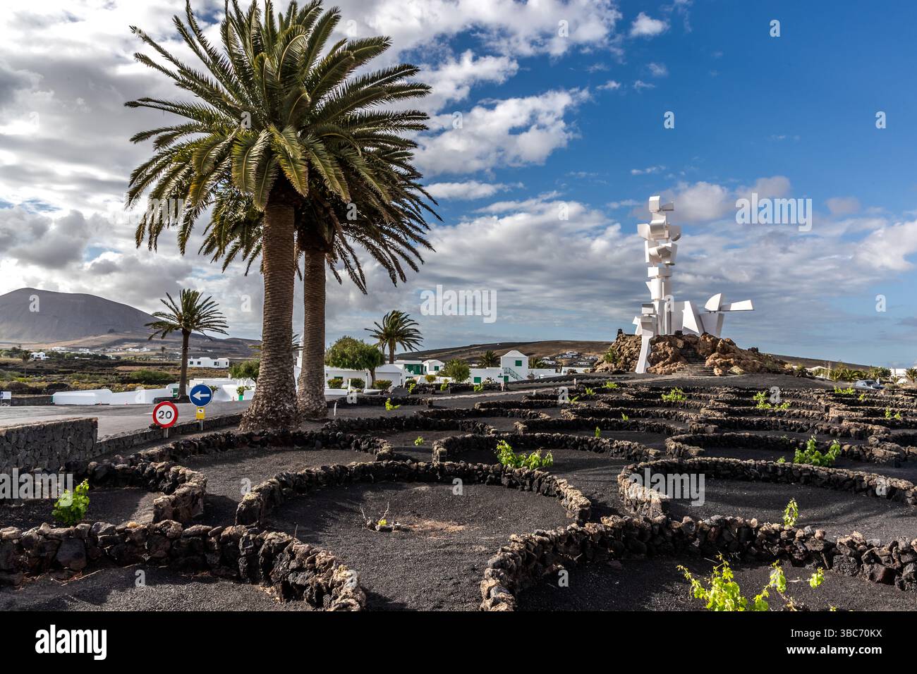 Circular vine craters walled with volcanic stone lie in the foreground near Mozaga on Lanzarote. The traditional technique shields individual vines from wind and drying out. The white Monumento al Campesino sculpture by César Manrique rises in the background. Carretera Arrecife a Tinajo, Mozaga, Canary Islands, Spain. Stock Photo