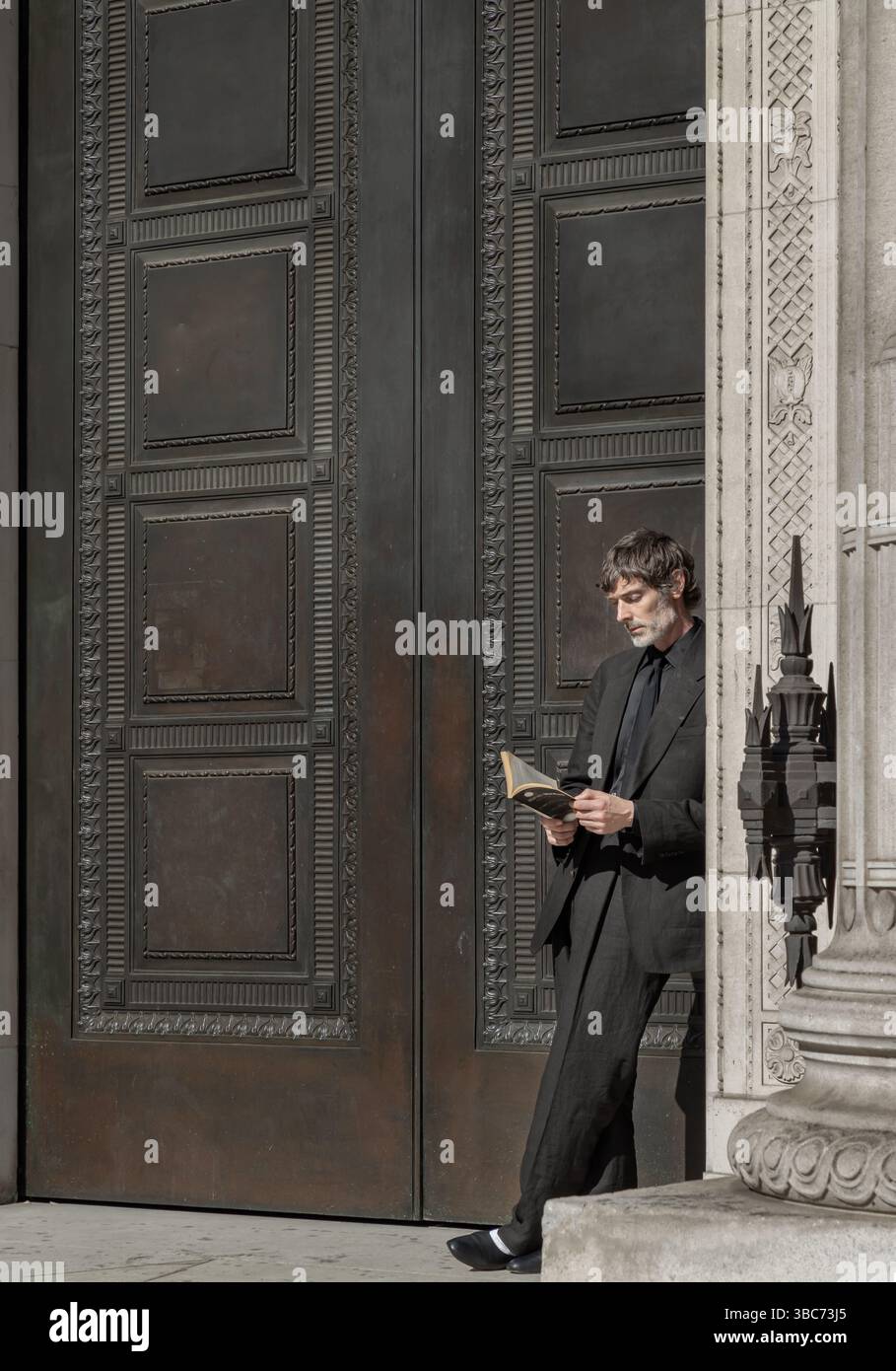 London, UK - May 13, 2025 - A man in suit standing in a doorway leaning against the door frame, reading a Haruki Murakami book. use it as your Wallpap Stock Photo