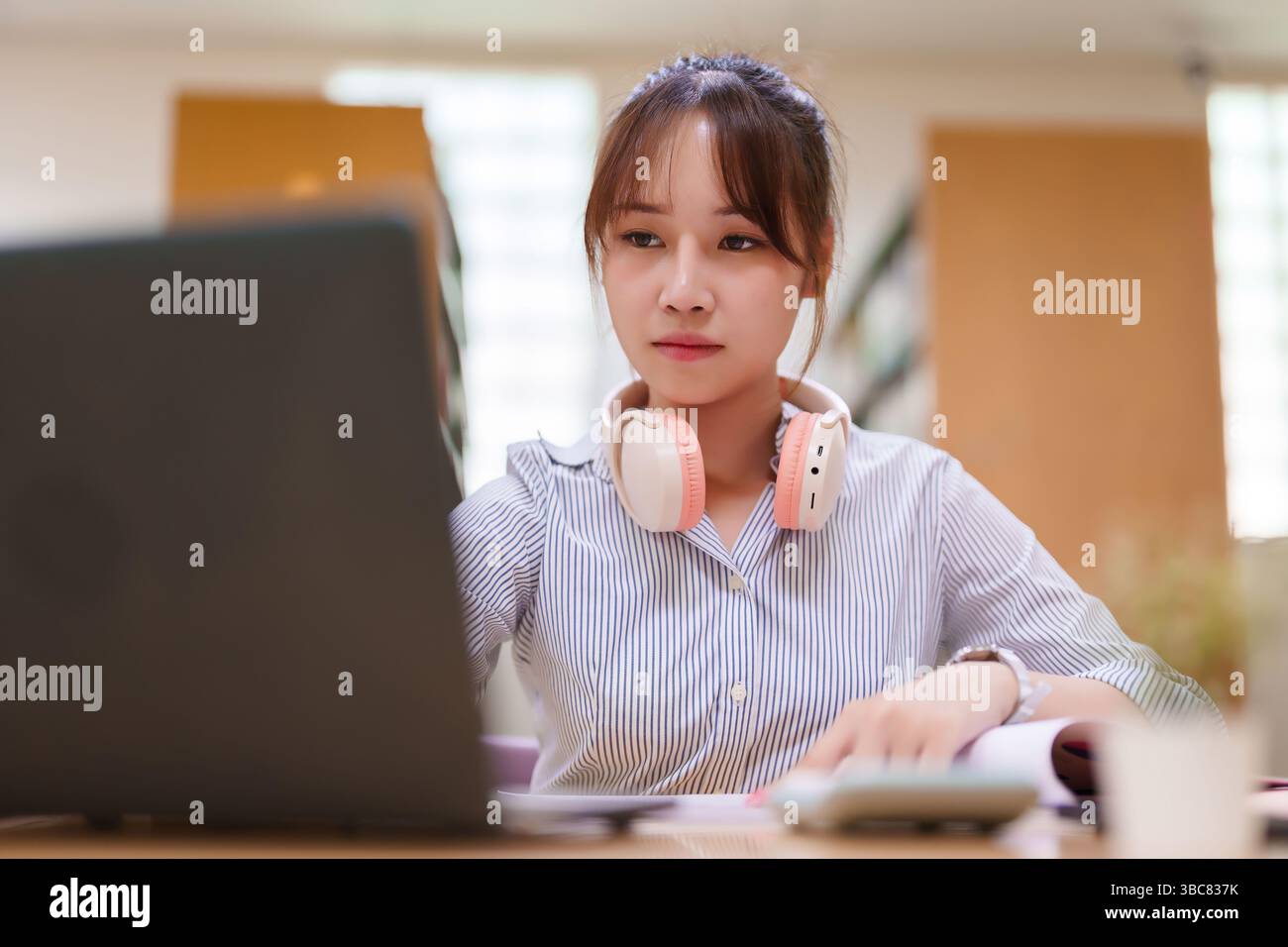 Image of a young Asian woman focused on her laptop, wearing headphones ...
