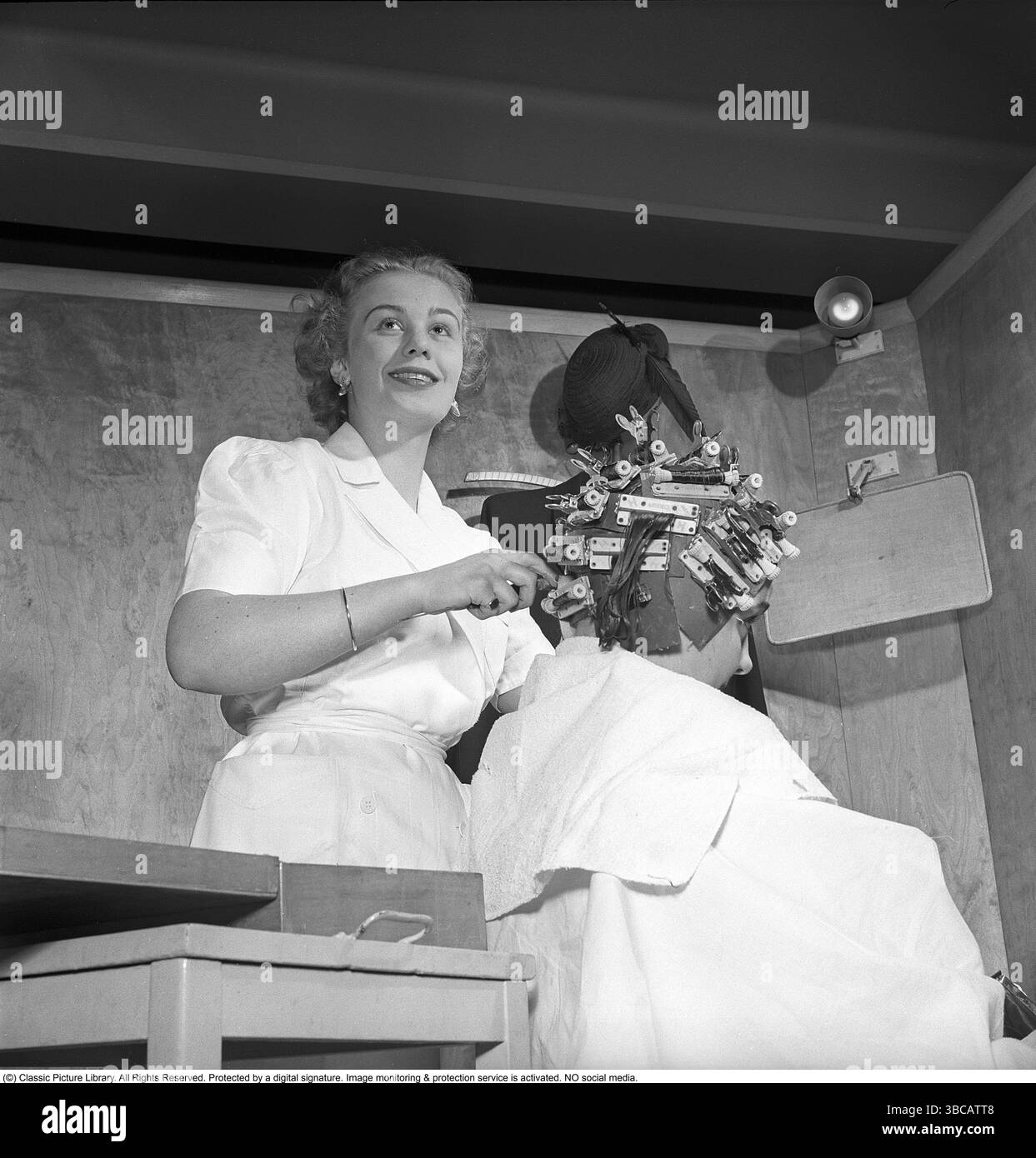 At the hairdresser 1955. A female customer is sitting in the chair at the salon presumably wishing for a curly hairdo as the hairdresser has put her hair in curlers, a lot of them. Stock Photo