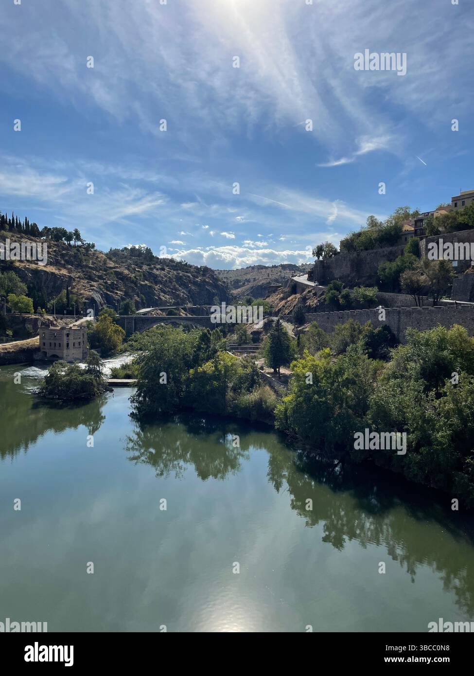 Scenic view of the Tagus River in Toledo, Spain, surrounded by lush trees, historic stone buildings, and hills under a bright blue sky. - Smartphone Captured Stock Image
