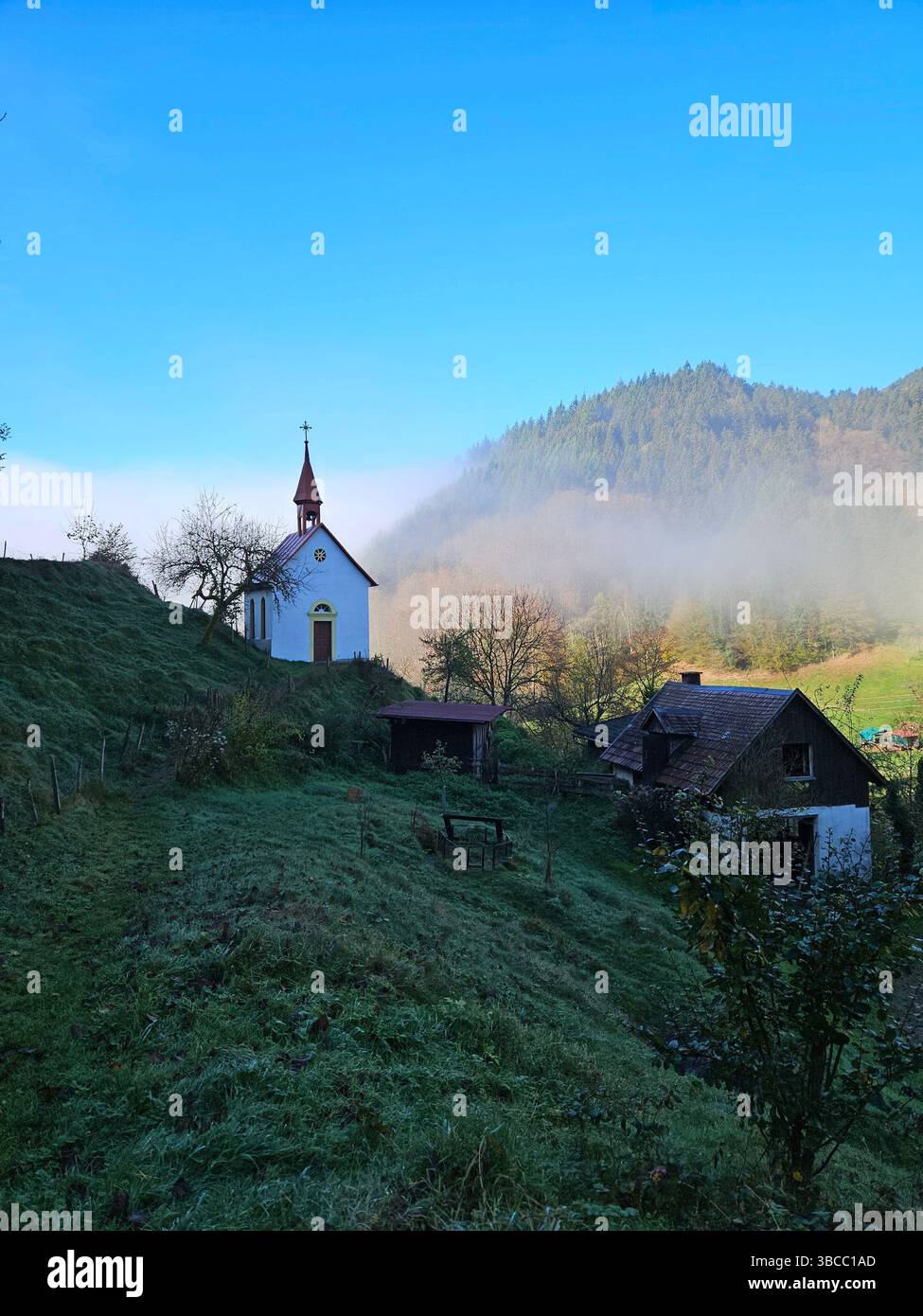 Misty autumn morning in the Black Forest, Germany. Hilltop chapel with rustic houses and colorful trees in peaceful rural mountain village. - Smartphone Captured Stock Image