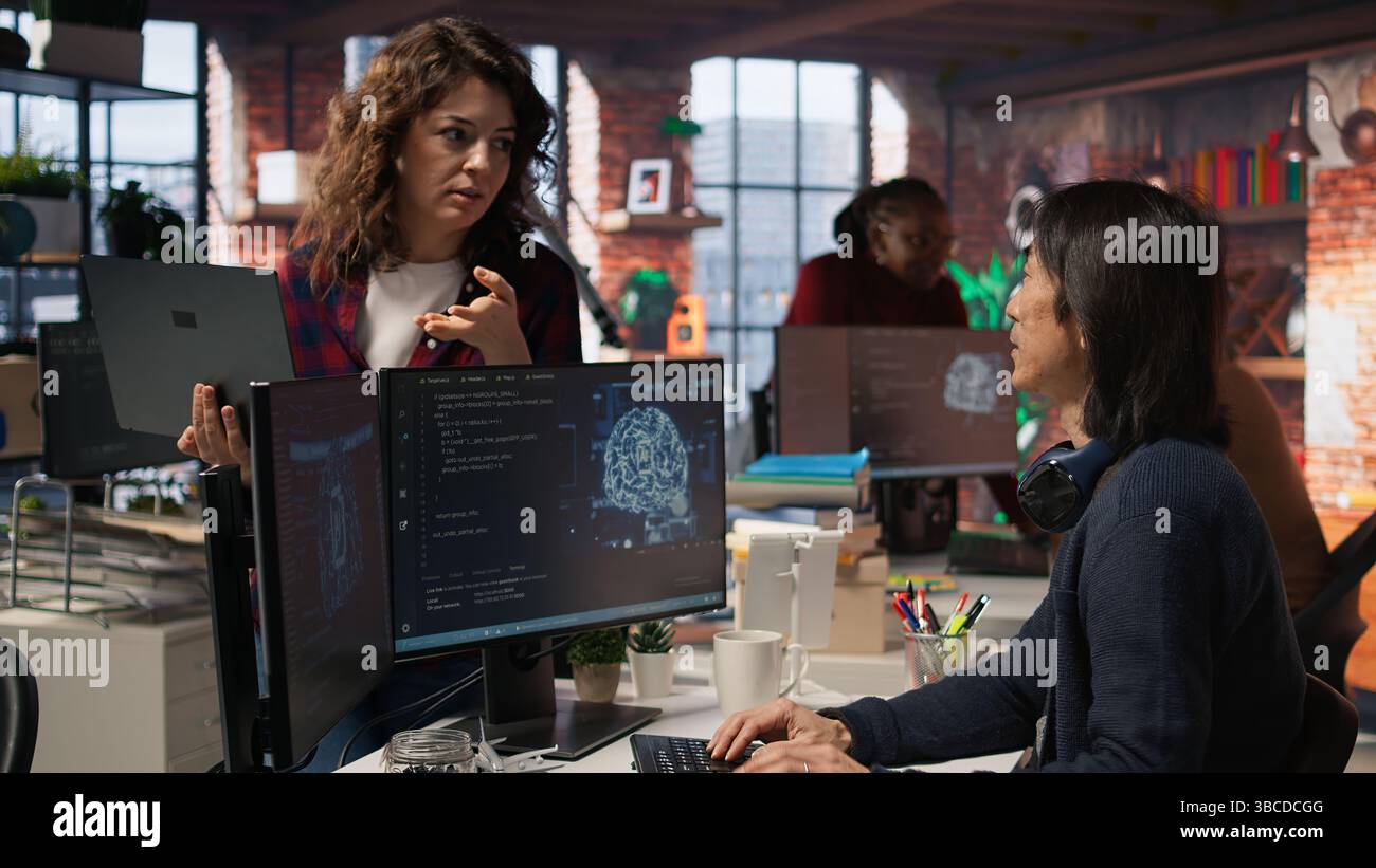 Startup intern holding laptop asks questions to senior technician coding advanced AI algorithm. Employee requests help from CTO manager in Silicon Valley workplace, camera A Stock Photo