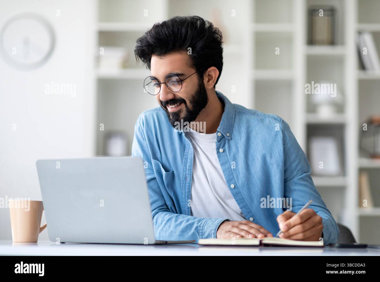 Online Lesson. Smiling Young Indian Man Using Laptop And Taking Notes Stock Photo