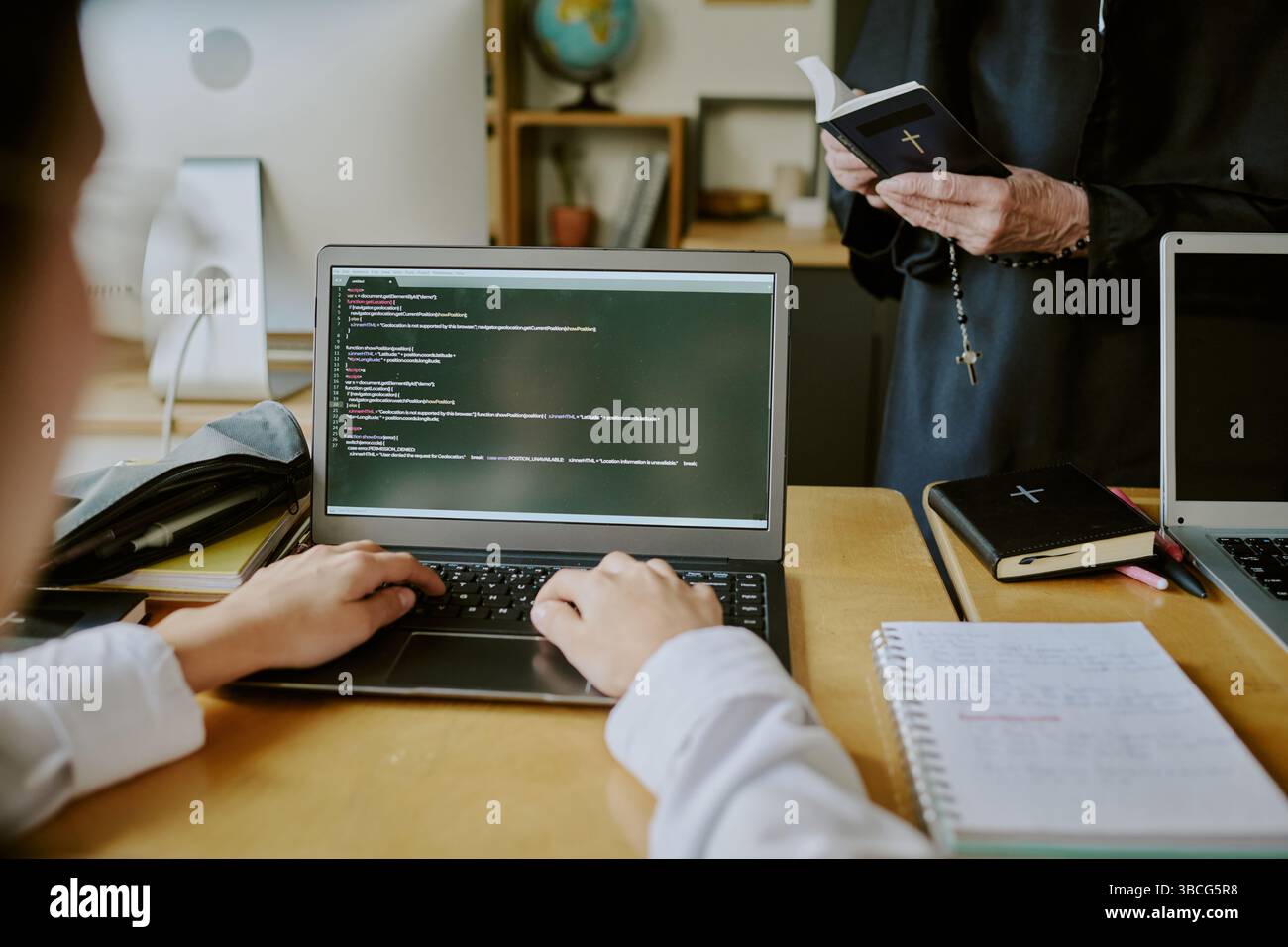 Students learning programming with a clergy member. Open laptops display code while notebooks lying on the desk, creating educational atmosphere Stock Photo