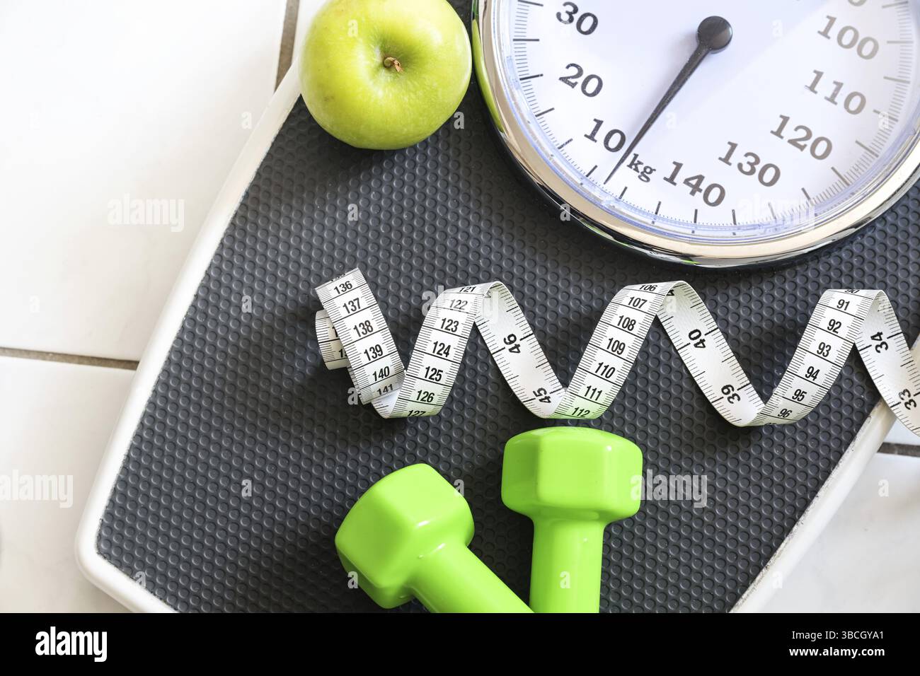 Part of a weight scale with measuring tape, green dumbbells and an apple on a tiled bathroom floor, fitness and slimming concept, top view from above Stock Photo