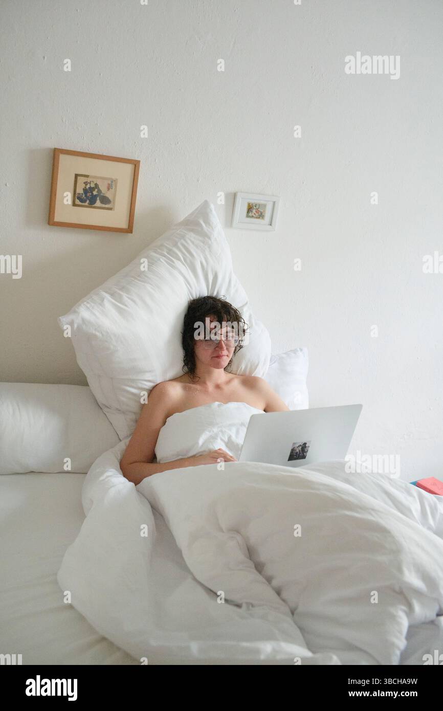 Young woman relaxing in white bedding while working on a laptop in a minimal bedroom. Stock Photo