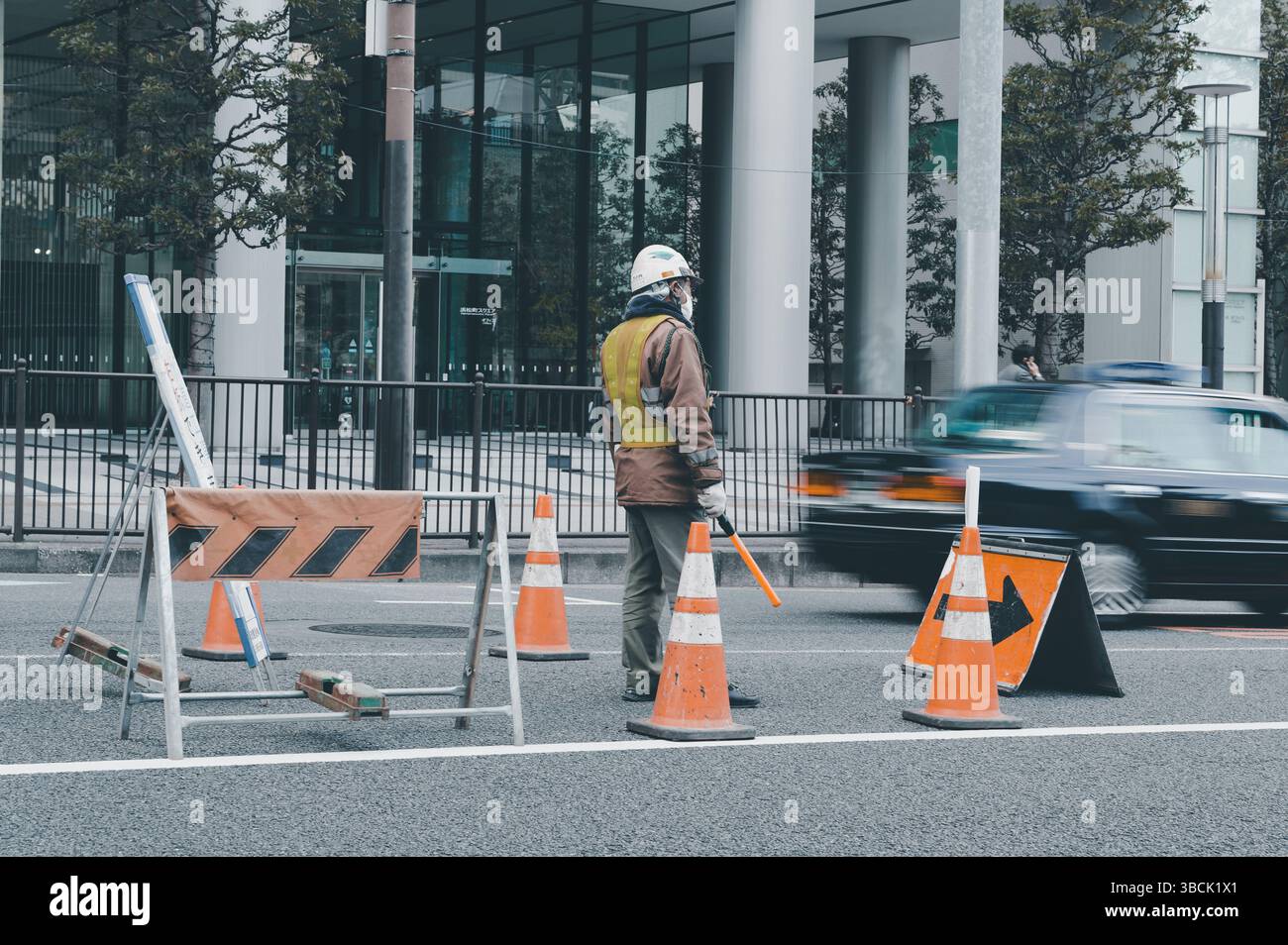 Traffic guard stands steady in central Tokyo as cars blur past a barricaded lane Stock Photo