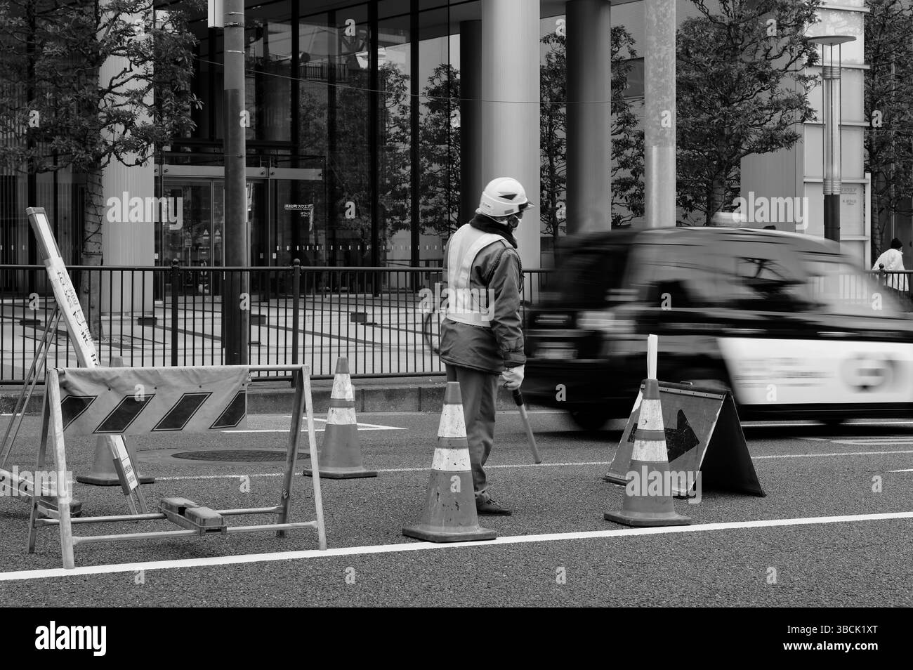 A road worker stands watch as a taxi rushes past, framed by cones and glass towers in monochrome Stock Photo