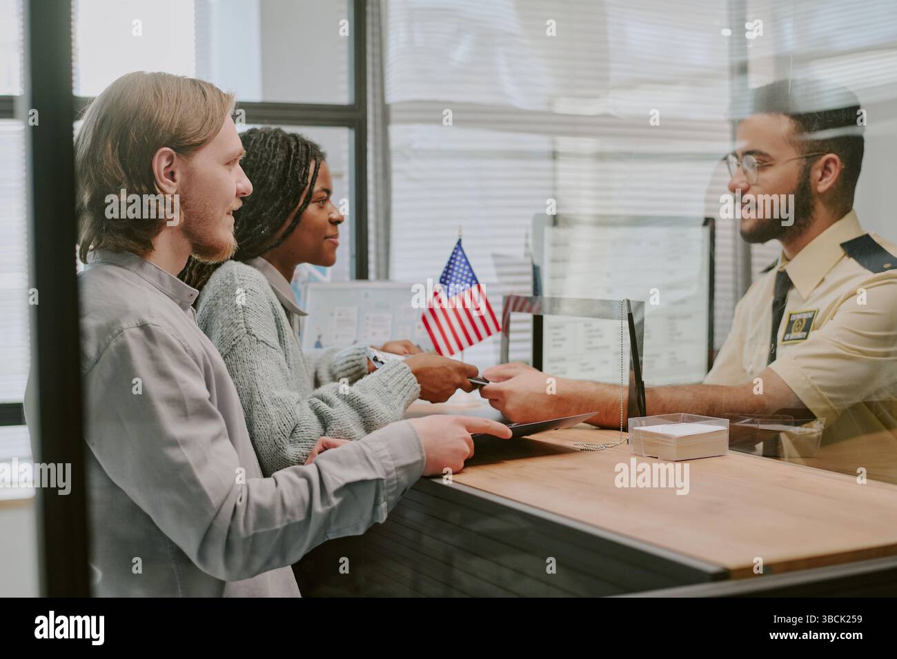 Diverse couple handing passports at immigration office counter with officer verifying documents and flag in background. Multi-ethnic group interacting Stock Photo
