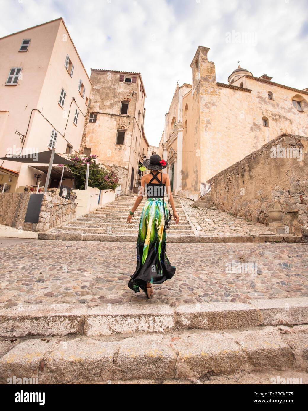 Fashionable woman is walking away from camera towards beautiful old stone city town Calvi in island Corsica, France. Model wears elegant outfit. Stock Photo