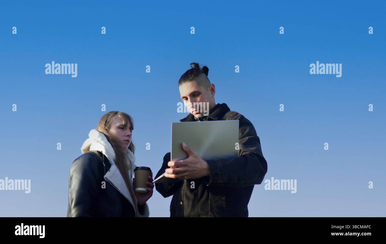 Two young professionals collaborating outdoors, using laptop and drinking coffee under blue sky Stock Photo