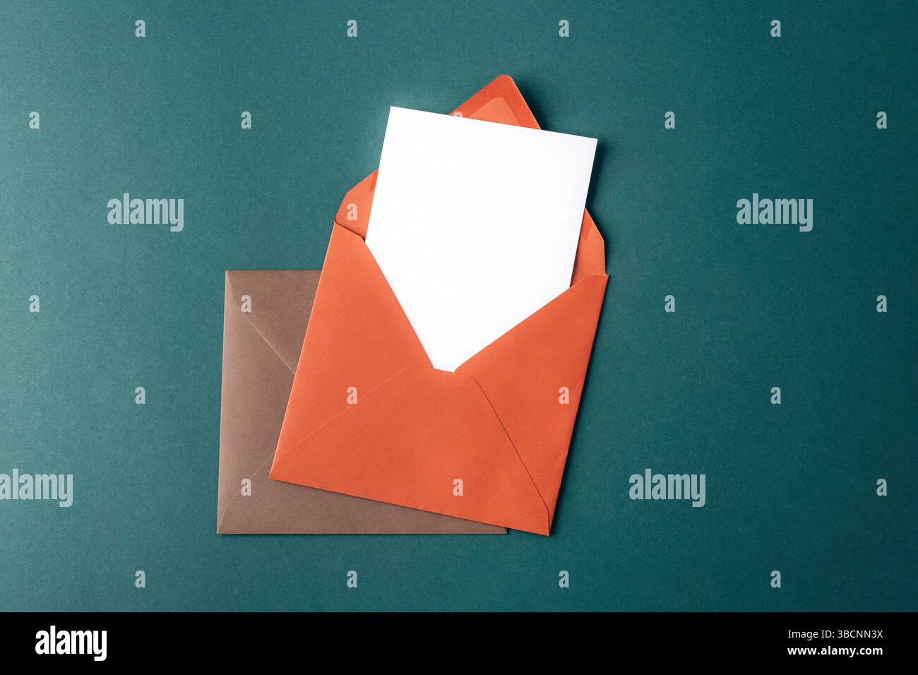 Orange and brown envelopes with blank white card on dark green background. Top view, flat lay, mockup. Stock Photo