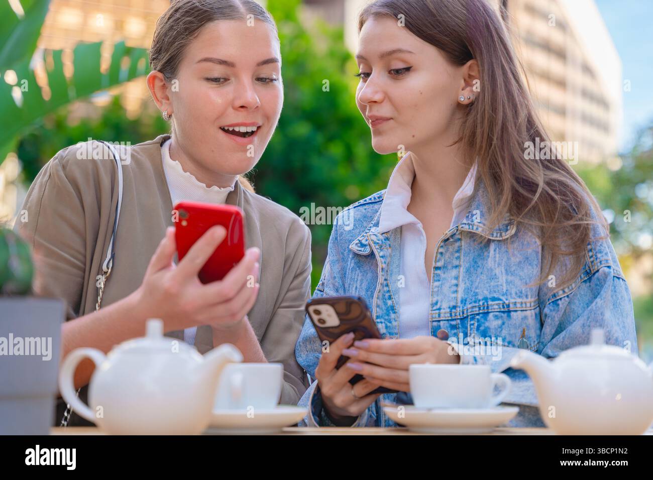 Two caucasian women using smartphones while enjoying tea at outdoor cafe table Stock Photo