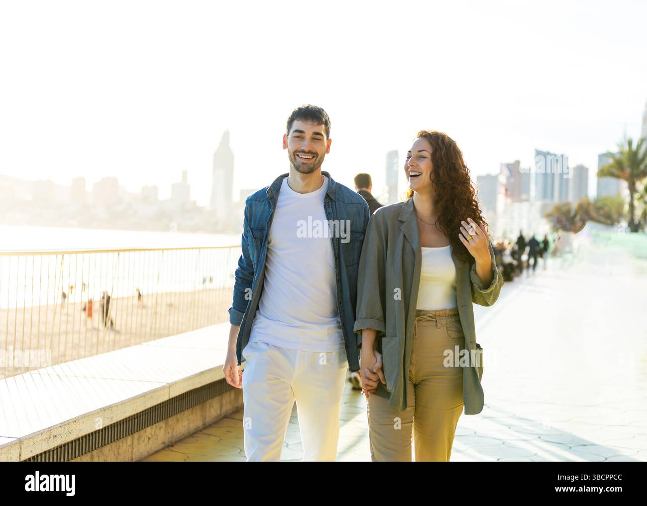 Young man and woman stroll together on a vibrant coastal walkway, holding hands and laughing under the bright sun with city skyline and beach in the b Stock Photo